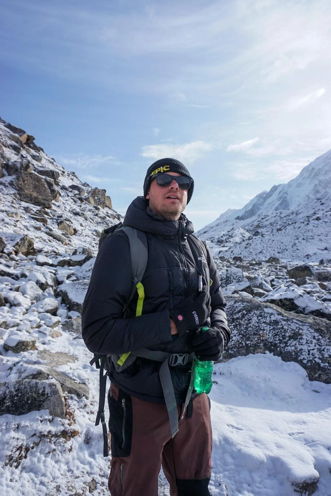 A man wearing sunglasses, a black beanie, and winter outdoor gear stands in a snow-covered mountain landscape, holding a green water bottle.
