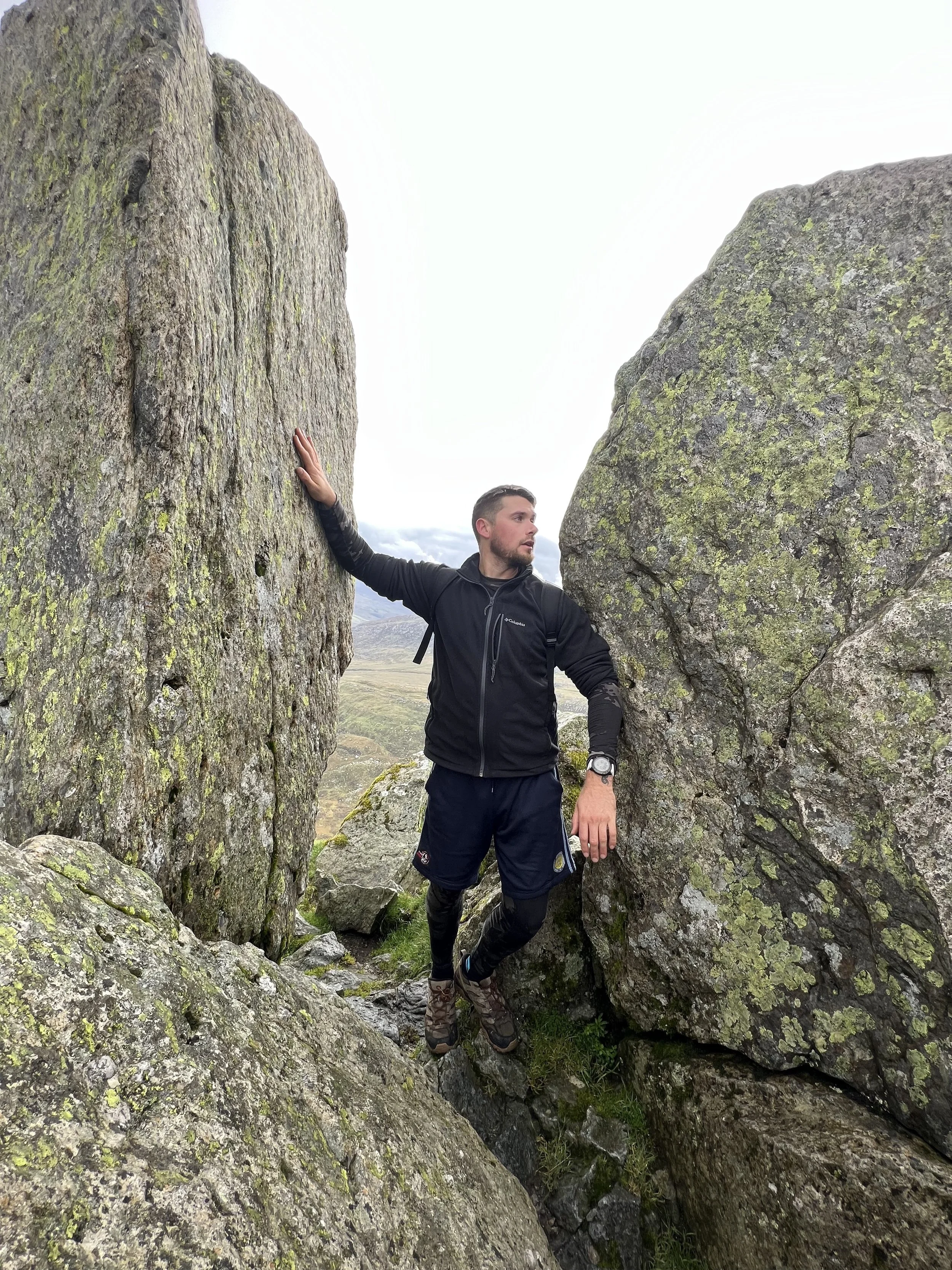 Man in black jacket and shorts standing between two large moss-covered rocks outdoors.