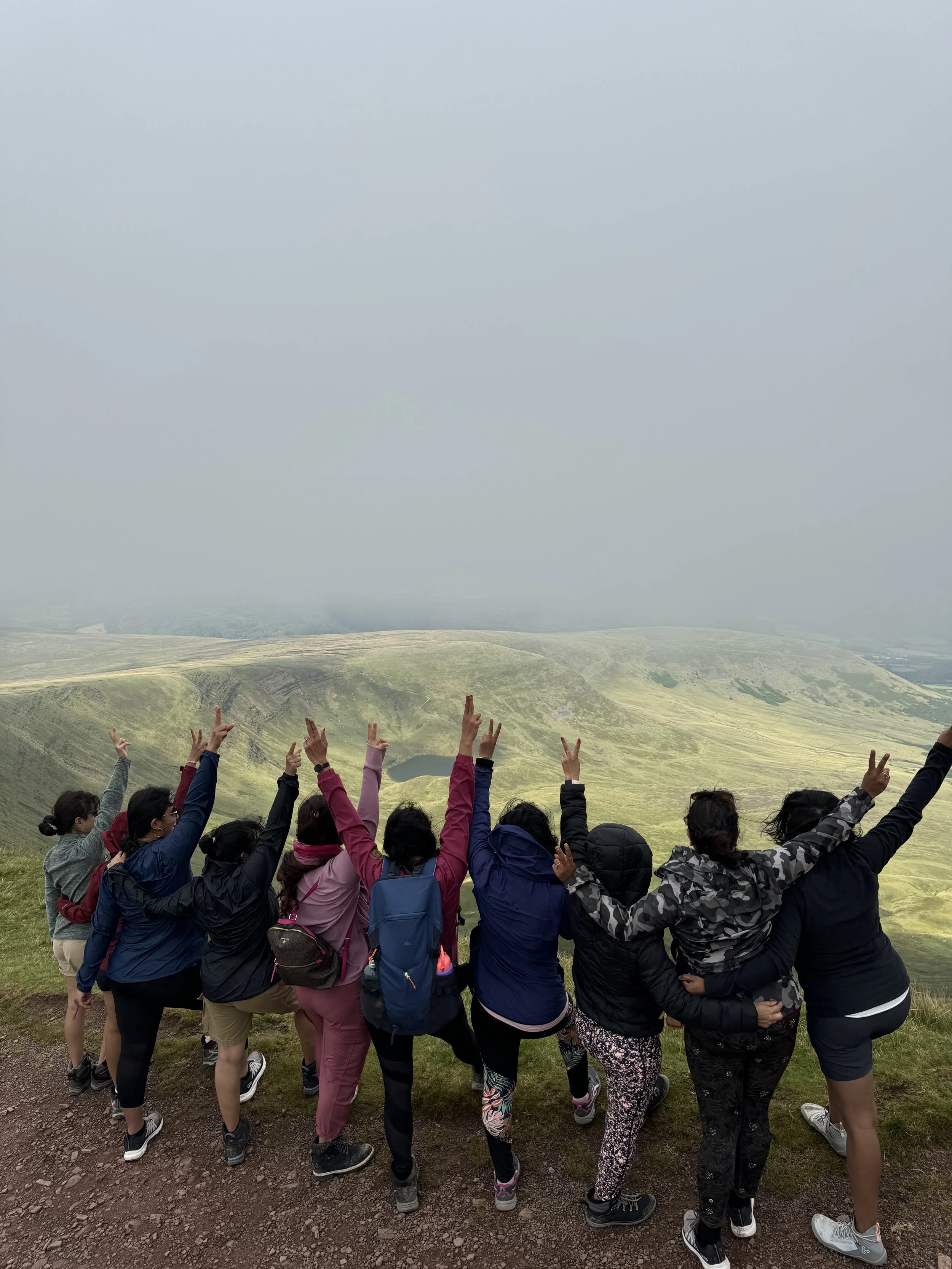Group of women hiking in outdoor landscape, posing with arms raised and peace signs, with rolling grassy hills and cloudy sky in background.
