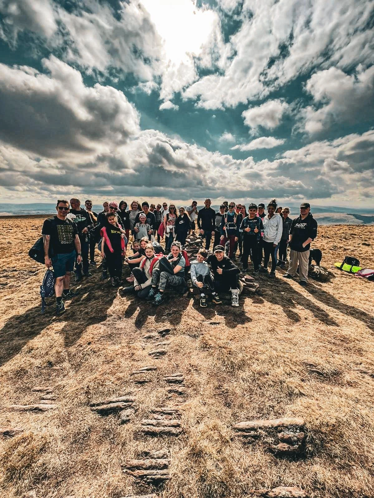 A large group of people, including children and adults, gathered on a hilltop with a scenic sky and cloudy sky in the background.
