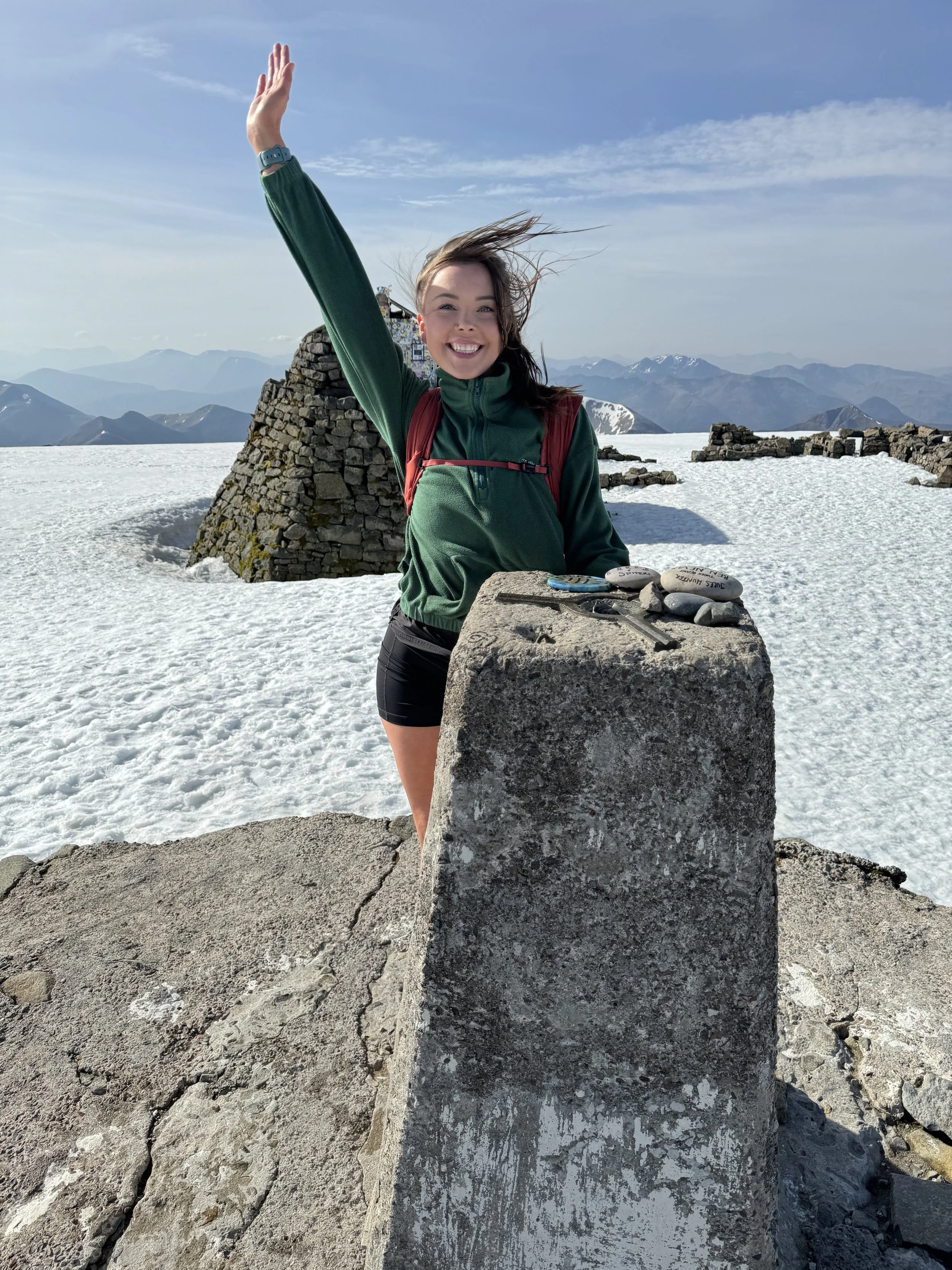 A woman with a green jacket and black shorts standing on snow-covered mountain top, smiling and waving, with a mountain landscape in the background.