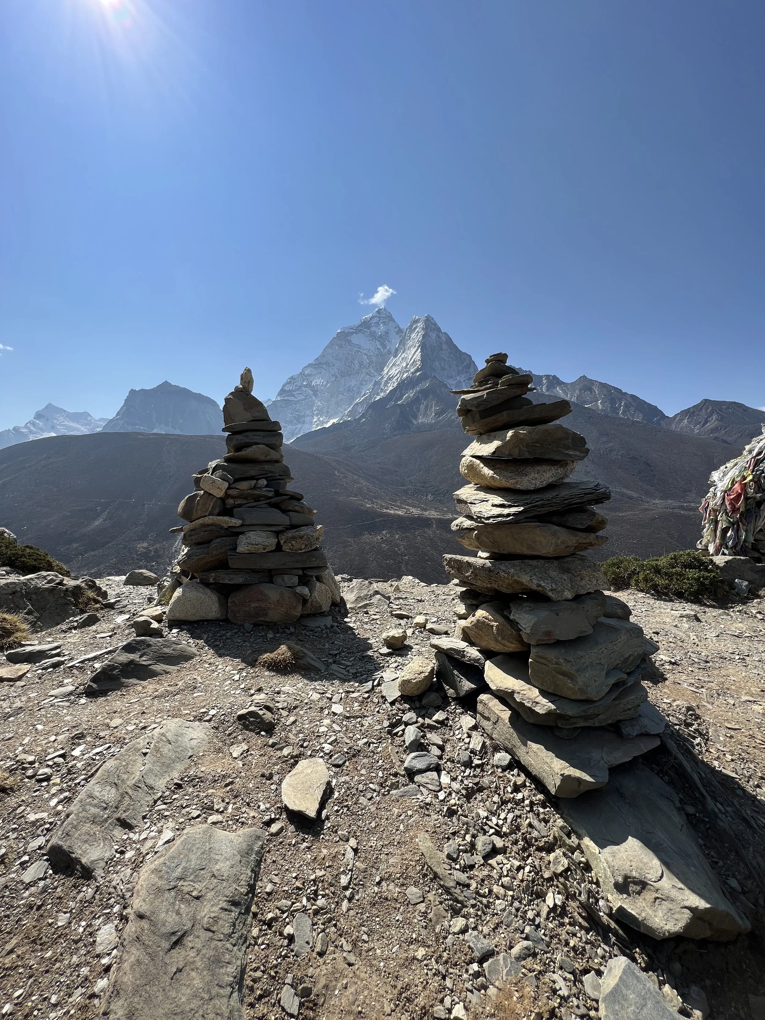 Two stacks of balanced rocks on rocky ground with mountain peaks in the background under a clear blue sky.