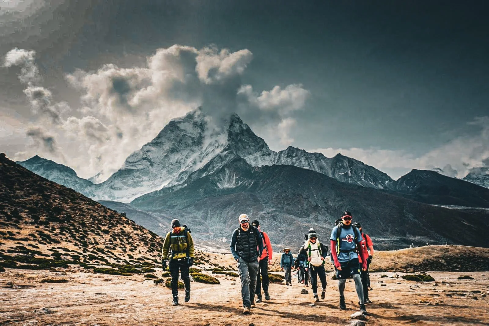 Group of hikers walking in mountainous terrain with snow-capped peaks and cloudy sky in the background.