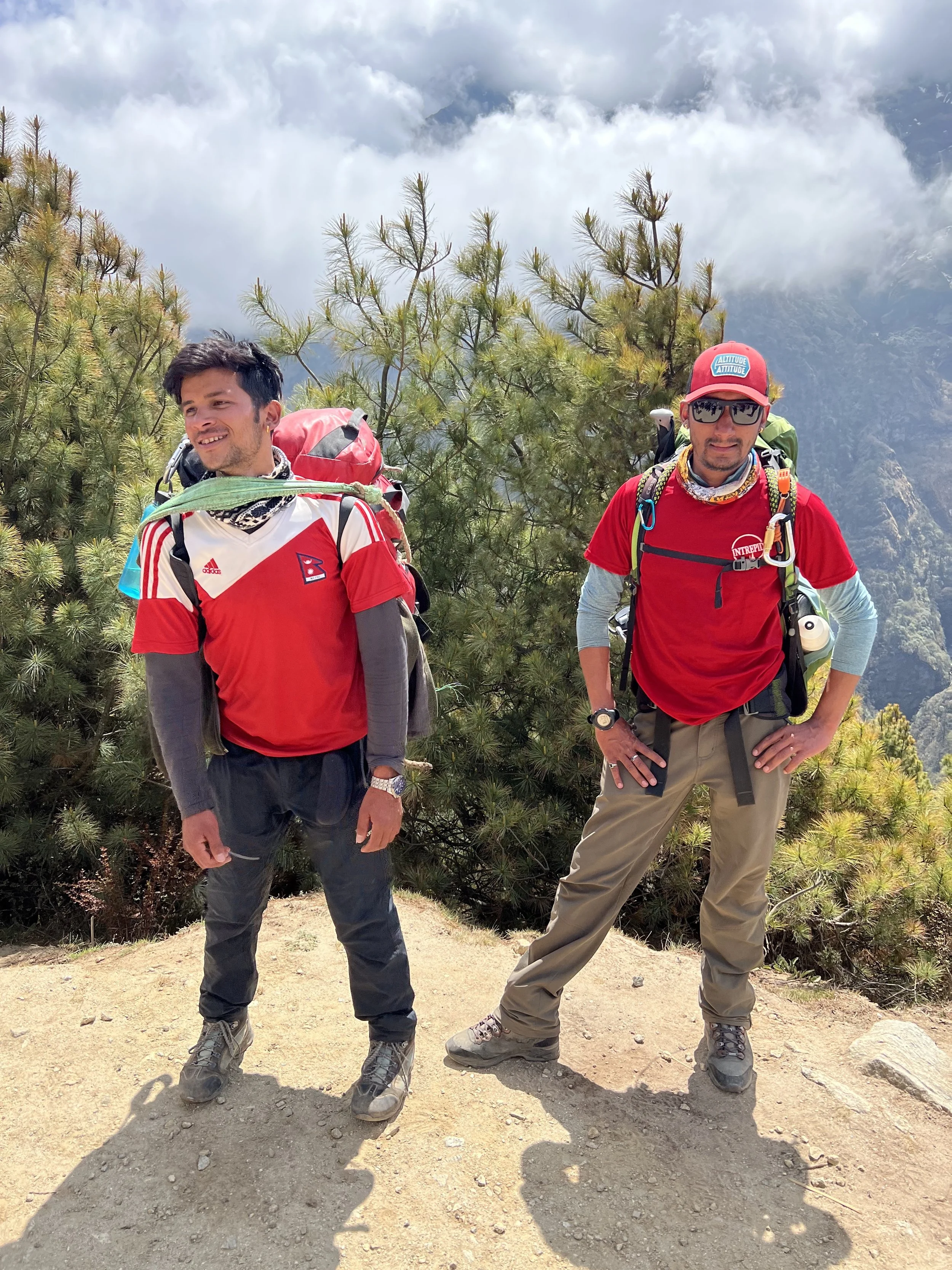 Two men hiking on a mountain trail, surrounded by pine trees and mountain scenery, wearing outdoor gear and backpacks.