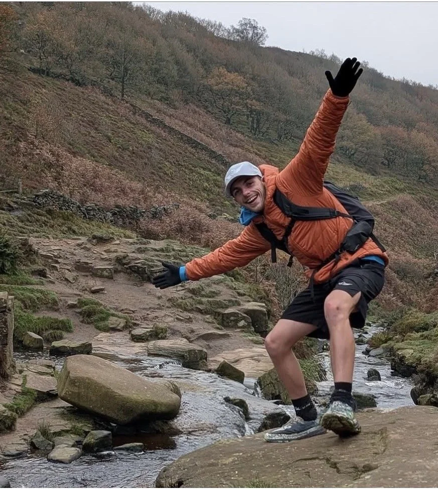 A young man in outdoor hiking gear balancing on rocks in a stream in a hilly, wooded area.