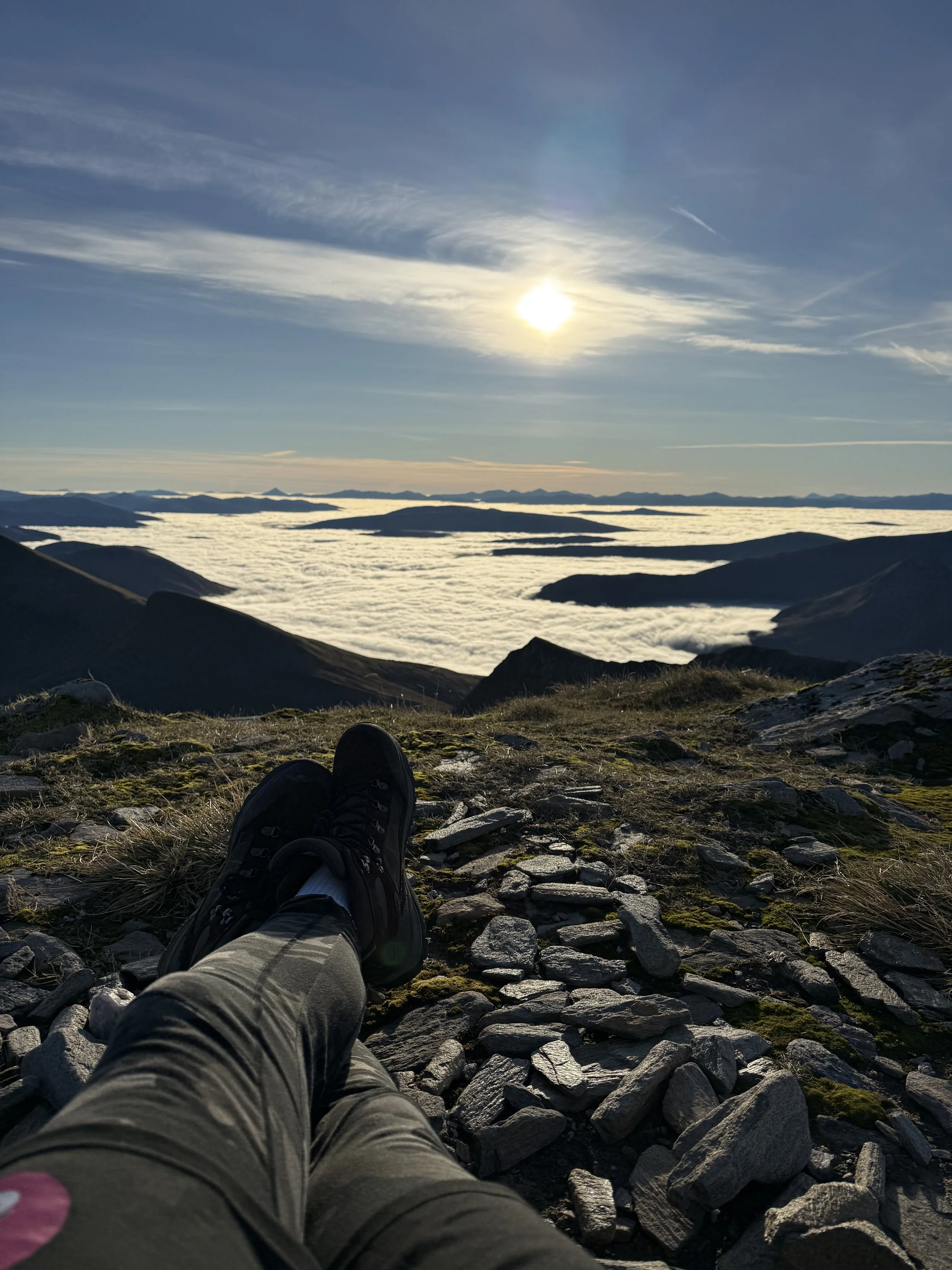 Person resting on a mountain with rock and grass, overlooking cloud-covered valleys, mountains in the distance, and a bright sun in a clear sky.