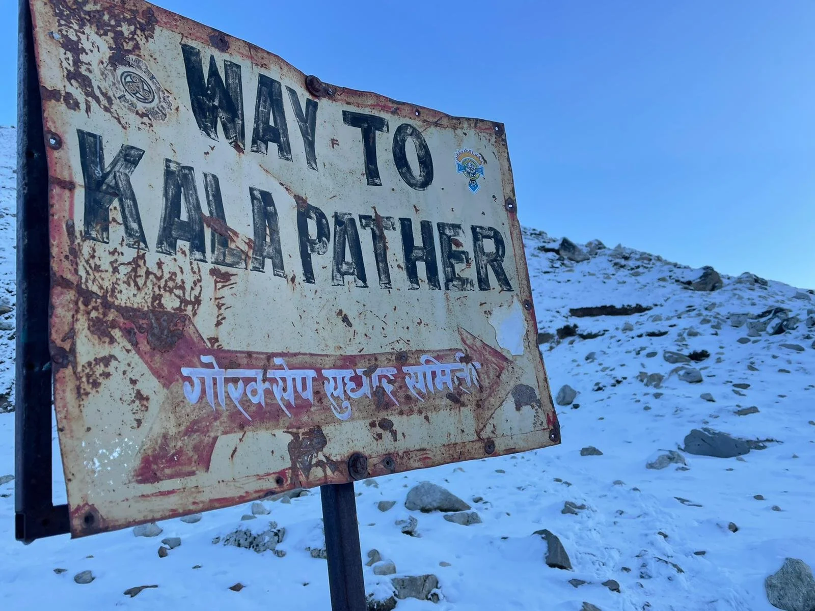 Rusty weathered sign with the text "Way to Khaar, Father" and text in another language, pointing toward a snowy mountainside.