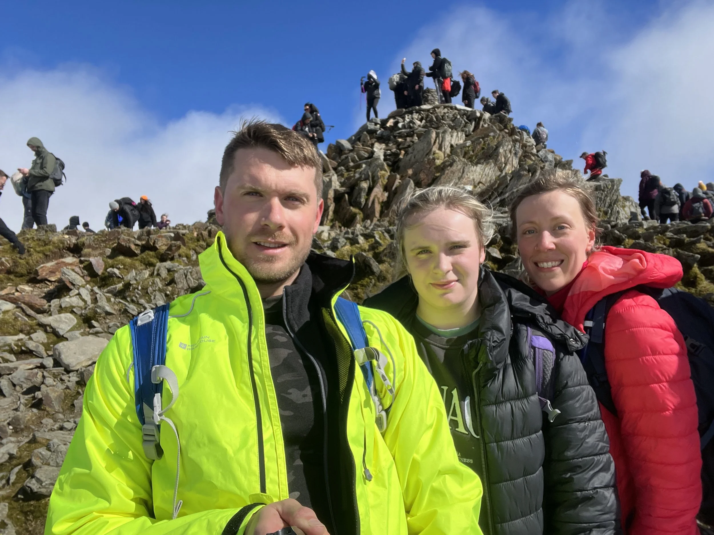 Three people standing on a mountain trail, smiling at the camera, with a rocky summit and other hikers in the background, dressed in outdoor gear.