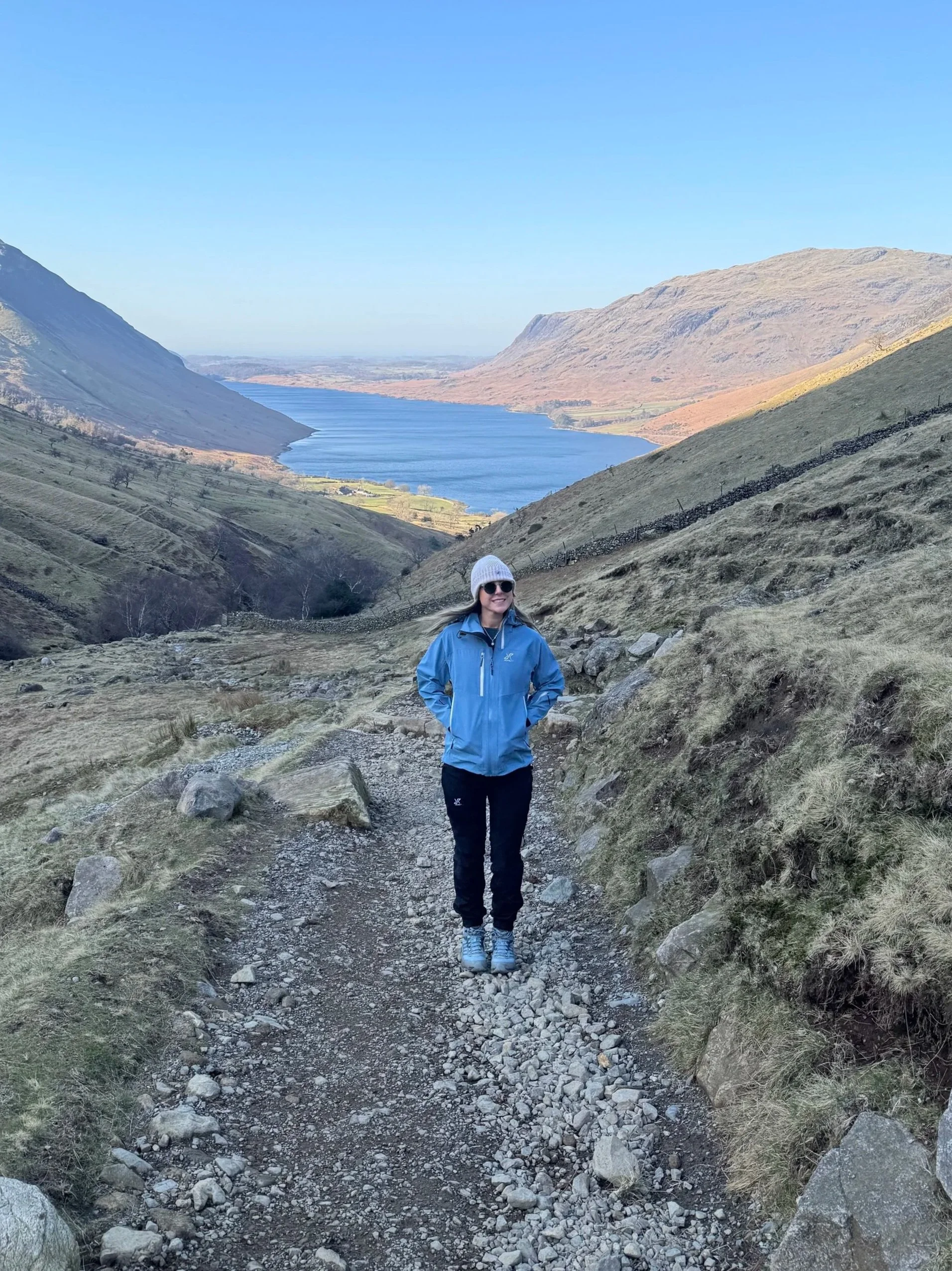 A woman in outdoor hiking gear walking on a rocky trail in a valley with hills, a lake, and mountains in the background under a clear blue sky.