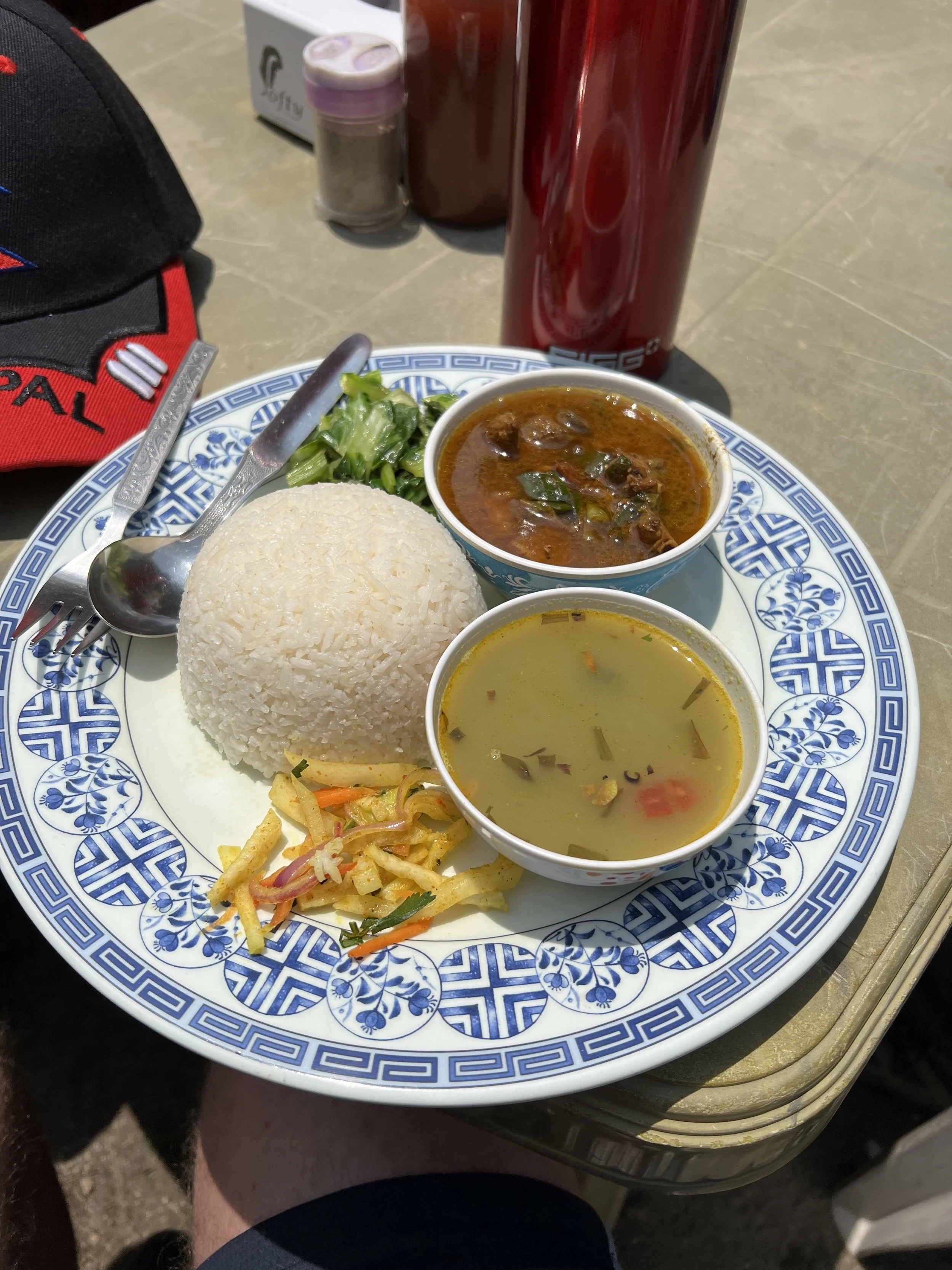 Plate of Indian food with rice, curry, soup, salad, and vegetable mix. Red thermos and condiments in the background.
