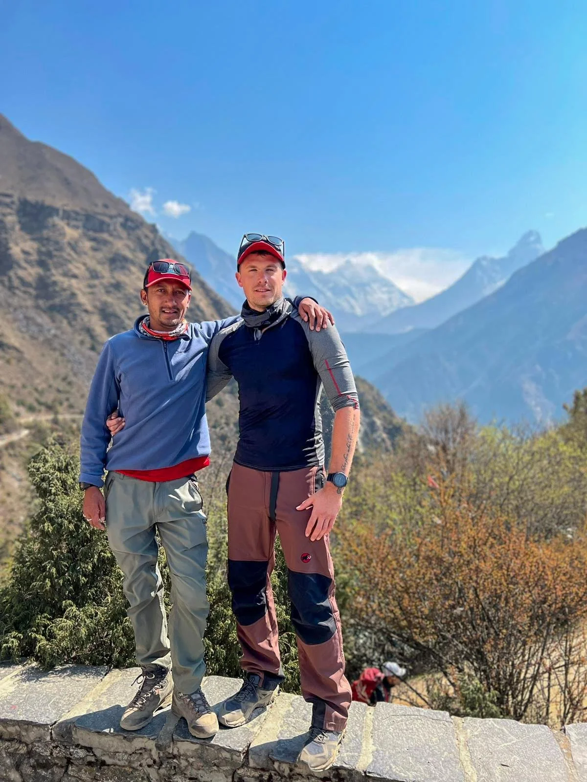 Two men standing on a rocky ledge with a mountainous landscape in the background. They are dressed in outdoor hiking gear and hats. One man has his arm around the other, and they are smiling at the camera.