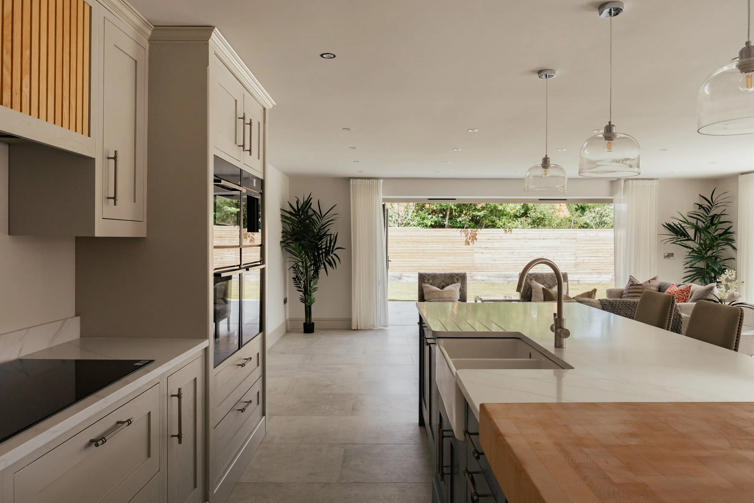 Modern kitchen with white cabinetry, a large island with a white sink, pendant lights hanging from the ceiling, and a view into a living area with large windows, green plants, and outdoor scenery.