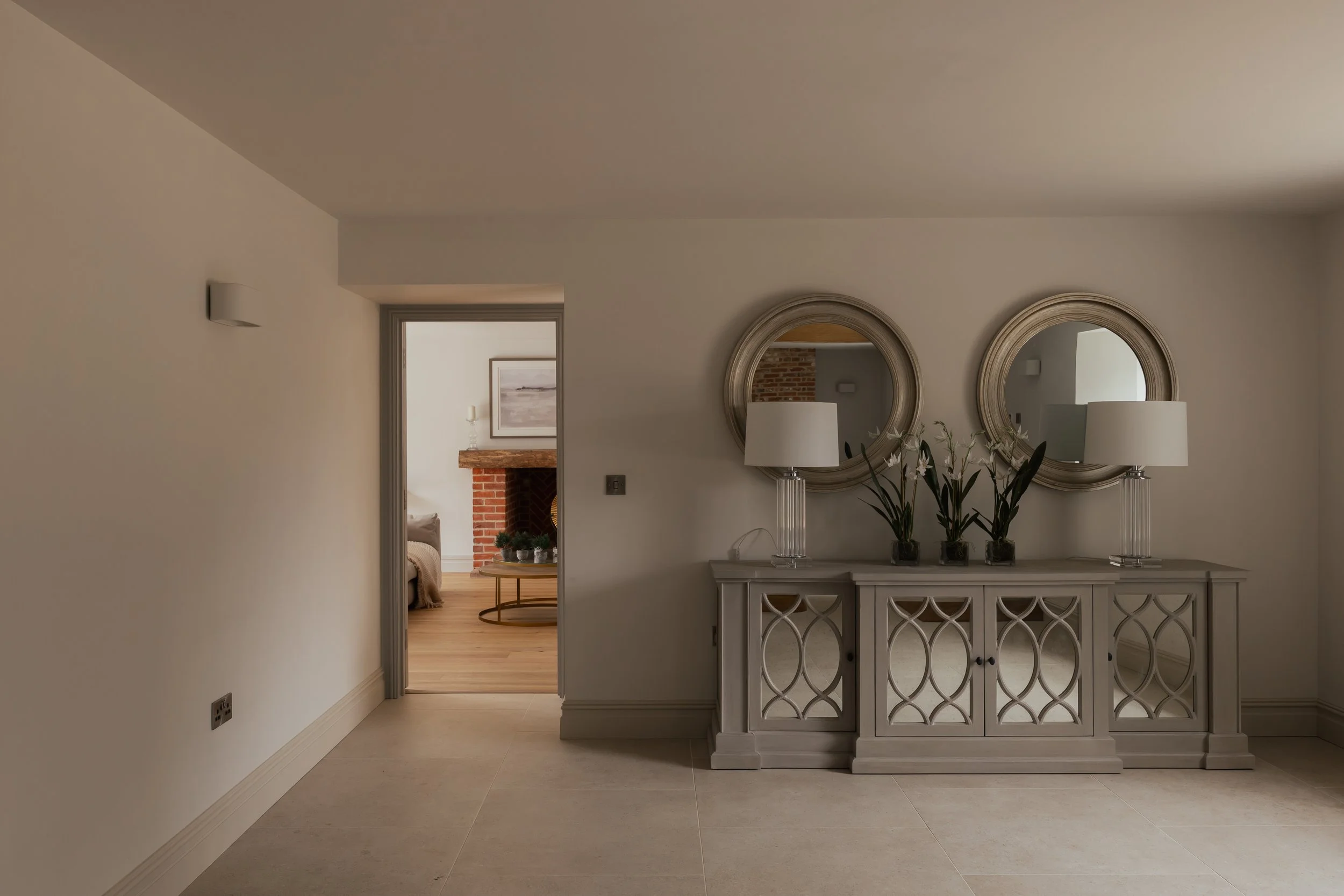 An elegant living room interior with a white sideboard decorated with three black vases and two white lamps, two round mirrors mounted above, and a hallway leading to a space with a brick fireplace and a round coffee table.