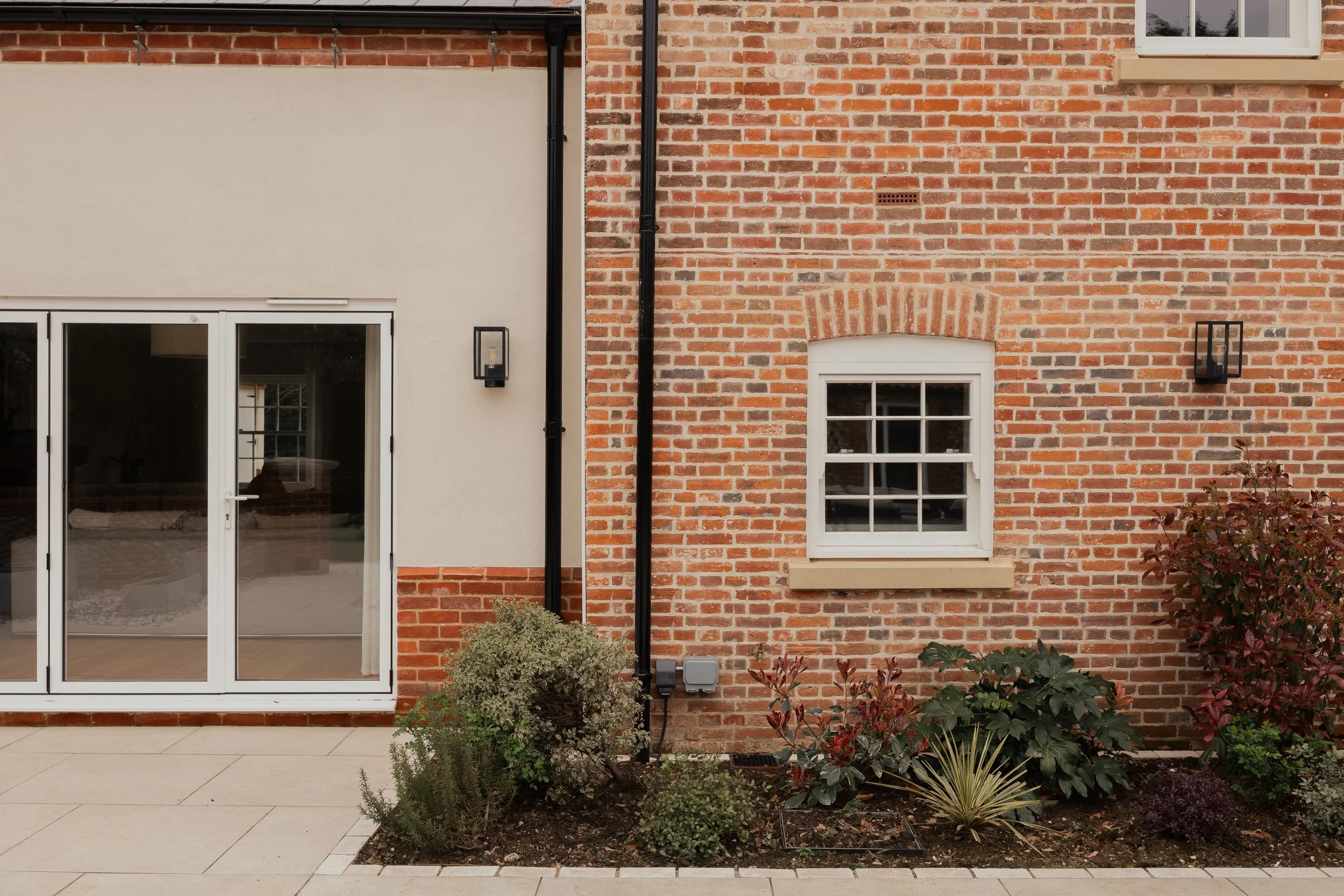 The image shows the exterior wall of a house with a mix of red brickwork and white painted wall sections. There are two black outdoor wall-mounted lantern lights, a balcony with glass sliding doors, and a window with white trim. The ground features beige tiles, and there are various small shrubs and plants in a garden bed in front of the wall.