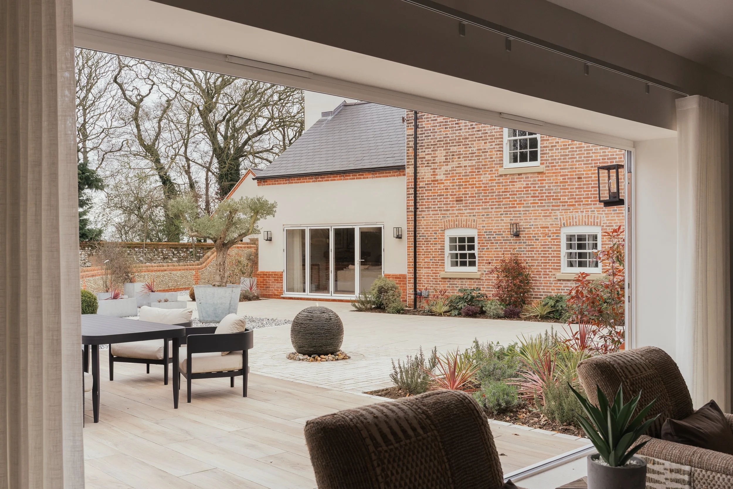 View of a modern townhouse backyard through a large window, showing an outdoor patio with seating, a decorative water fountain, landscaped plants, and a brick and white facade house with large sliding glass doors.