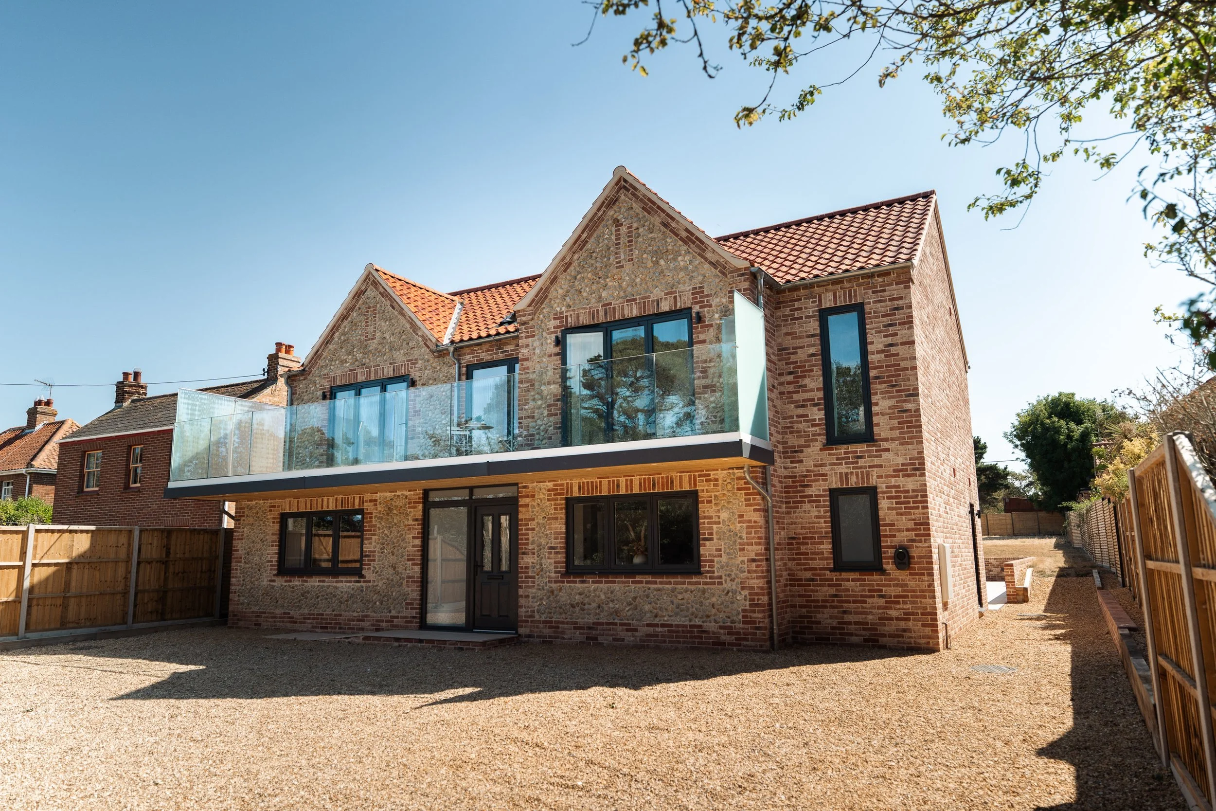 A modern brick house with large windows and a glass balcony, situated in a sunny backyard with a gravel ground and surrounded by a wooden fence.