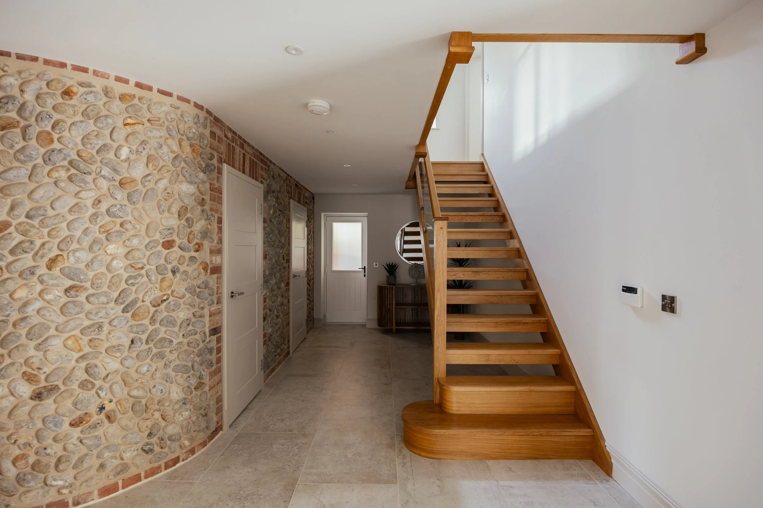 Interior view of a modern house with a wooden staircase, white and brick stone walls, a small mirror, and a door with frosted glass.