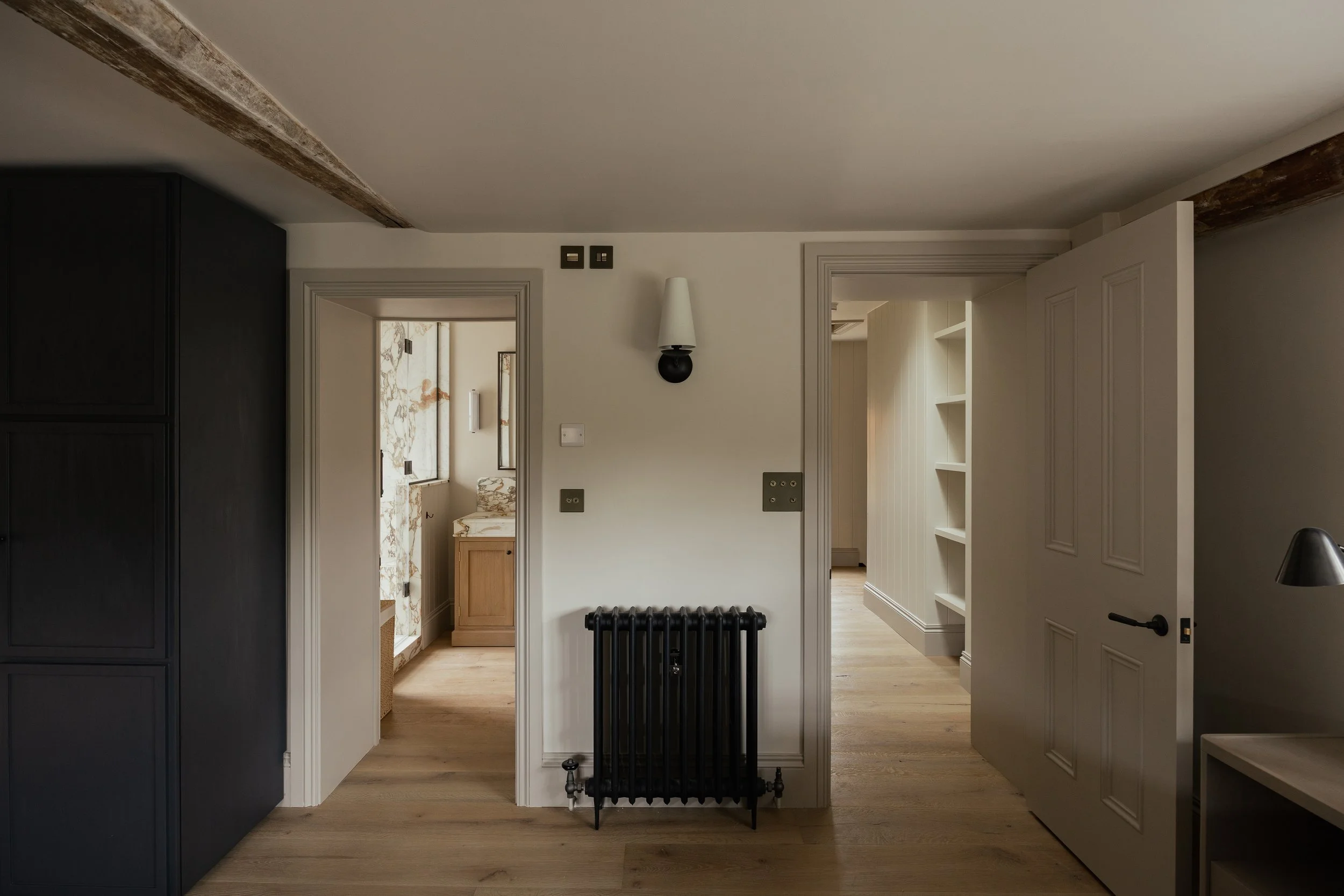 Interior view of a room with white walls, wooden flooring, and visible doorway leading to a bathroom with marble tiles and a small wooden cabinet. There is a black radiator and a wall-mounted light fixture, and parts of other rooms are visible.