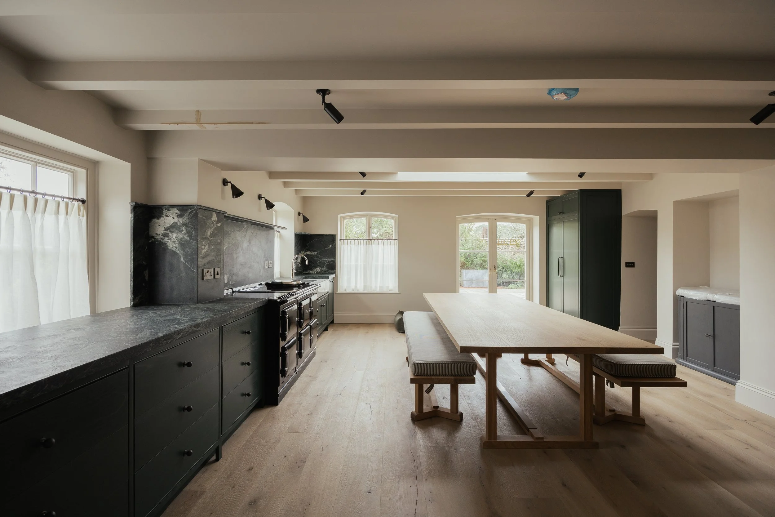 Open-concept kitchen and dining area with black cabinetry, marble countertops, wooden dining table, light-colored hardwood floors, large windows with white curtains, and recessed ceiling lighting.