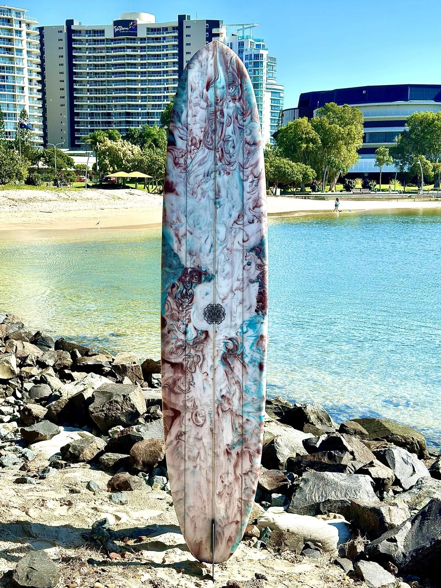 Marbled surfboard standing upright on rocks near a beach with trees and high-rise buildings in the background.