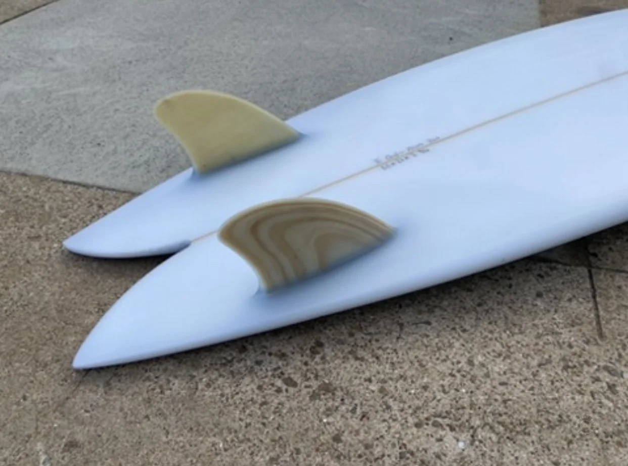 Two white surfboards lying on a sandy and concrete surface, with yellowish fins attached, seen from a side angle.