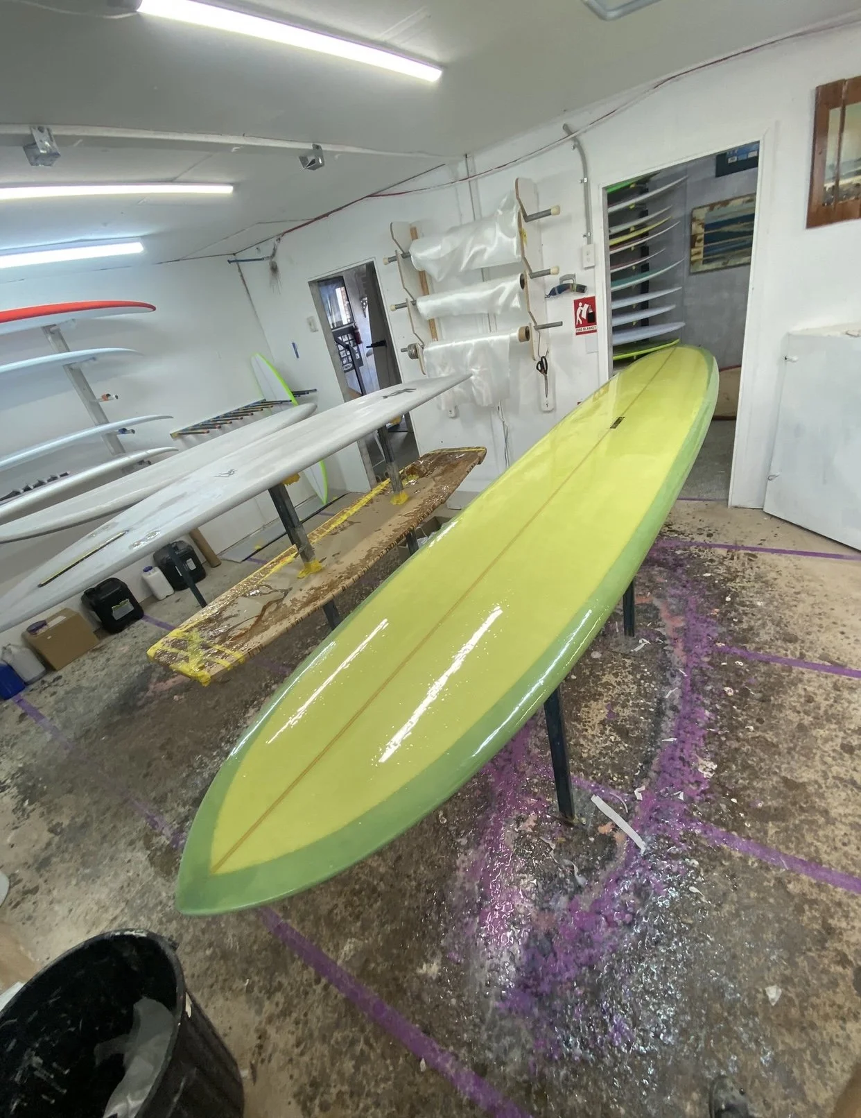 A surfboard shaping workshop, with a yellow surfboard on stands, other surfboards hanging on the wall, and various tools and materials around the workshop.
