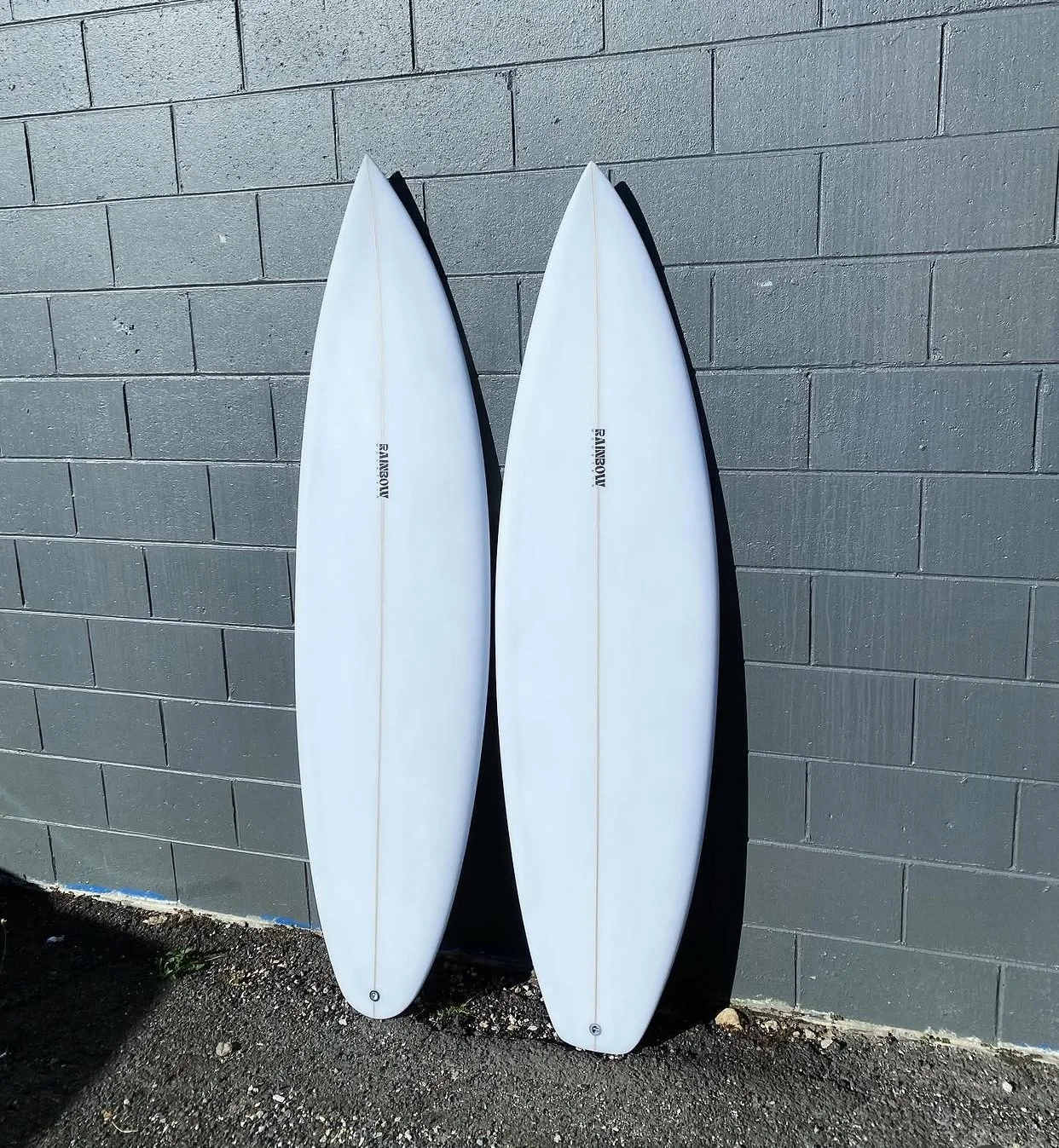 Two white surfboards leaning against a gray brick wall outdoors on a sunny day.