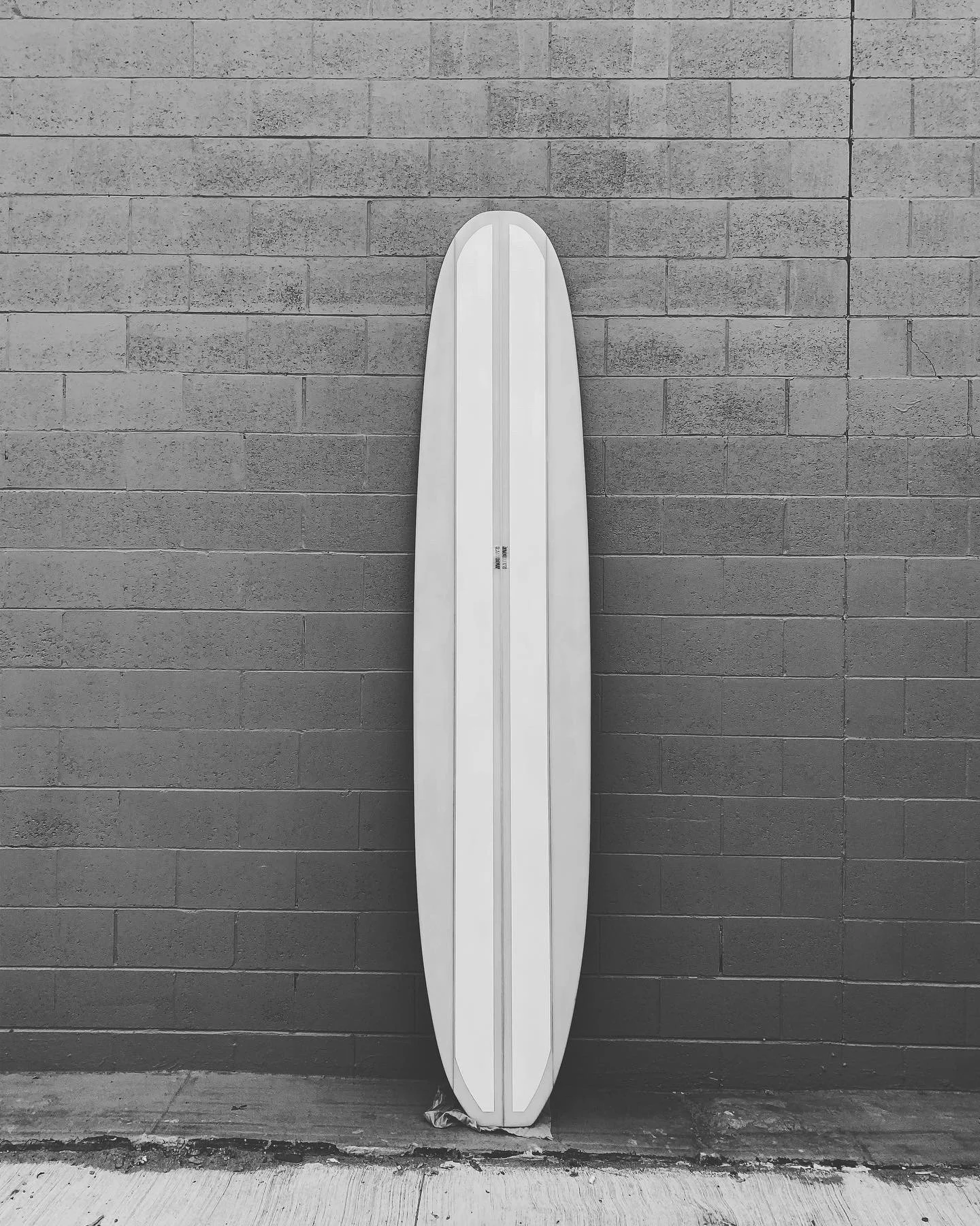 A white surfboard leaning against a gray brick wall outside on a sidewalk.
