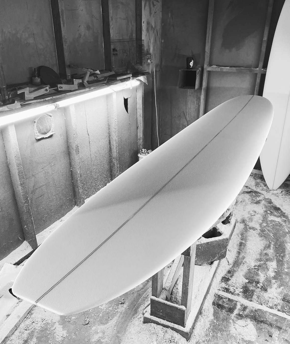 A surfboard with a stringer, resting on a stand in a workshop with unfinished walls and a workbench.
