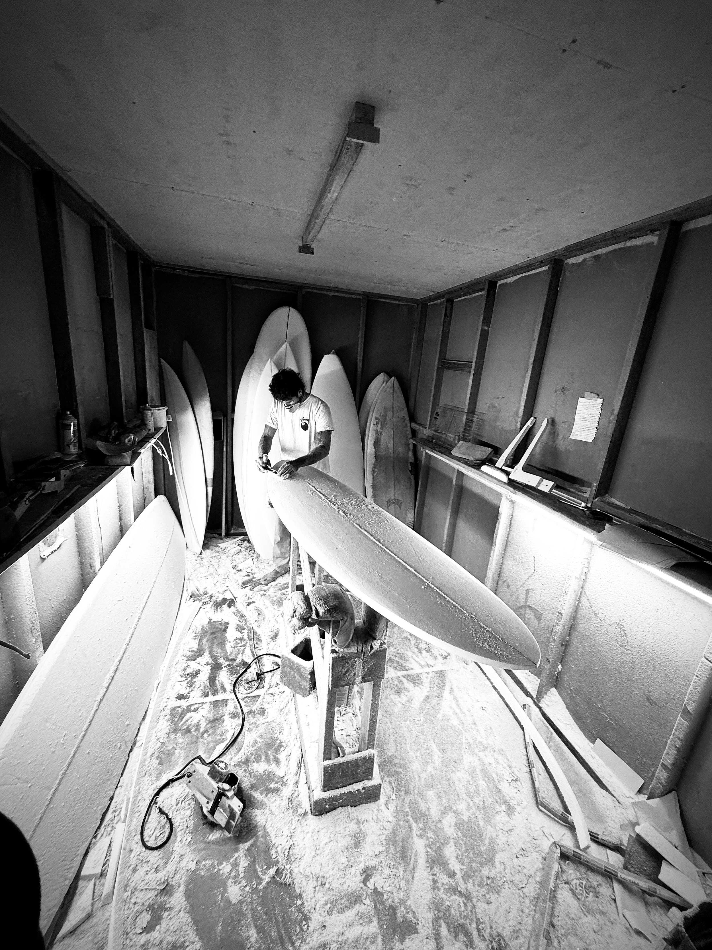 A person working on a surfboard in a workshop, surrounded by tools and unfinished boards, with a workbench and shelves on the side.