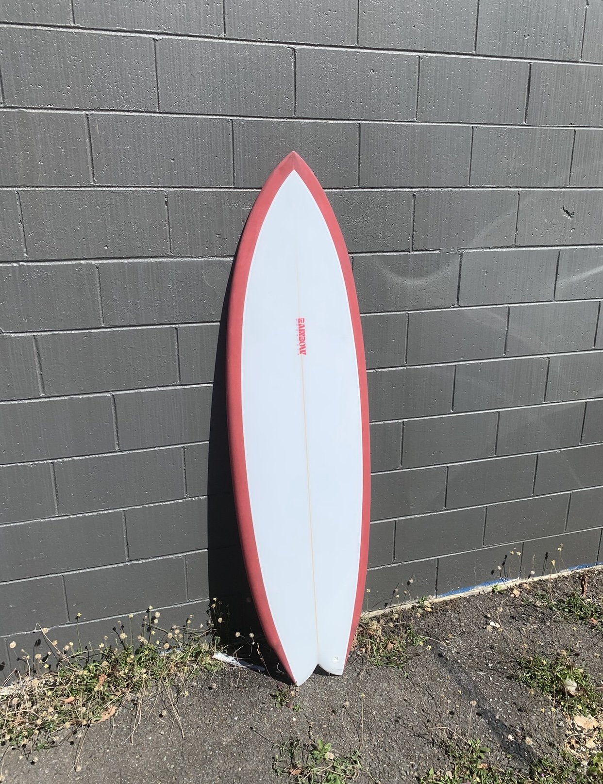 White surfboard with red border leaning against a gray brick wall outdoors