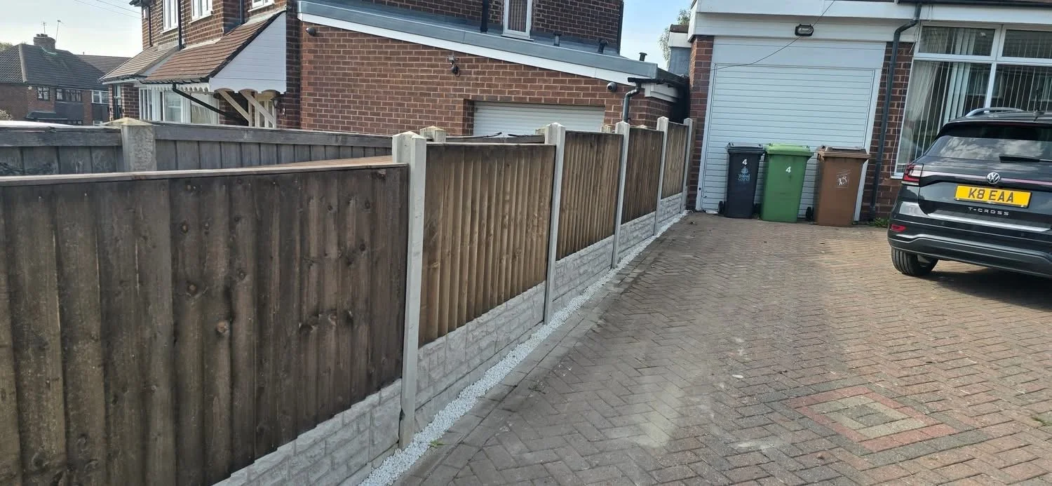 View of a paved driveway next to a wooden and concrete fence, with a house and several trash bins in the background, and a black Volkswagen SUV parked on the right.