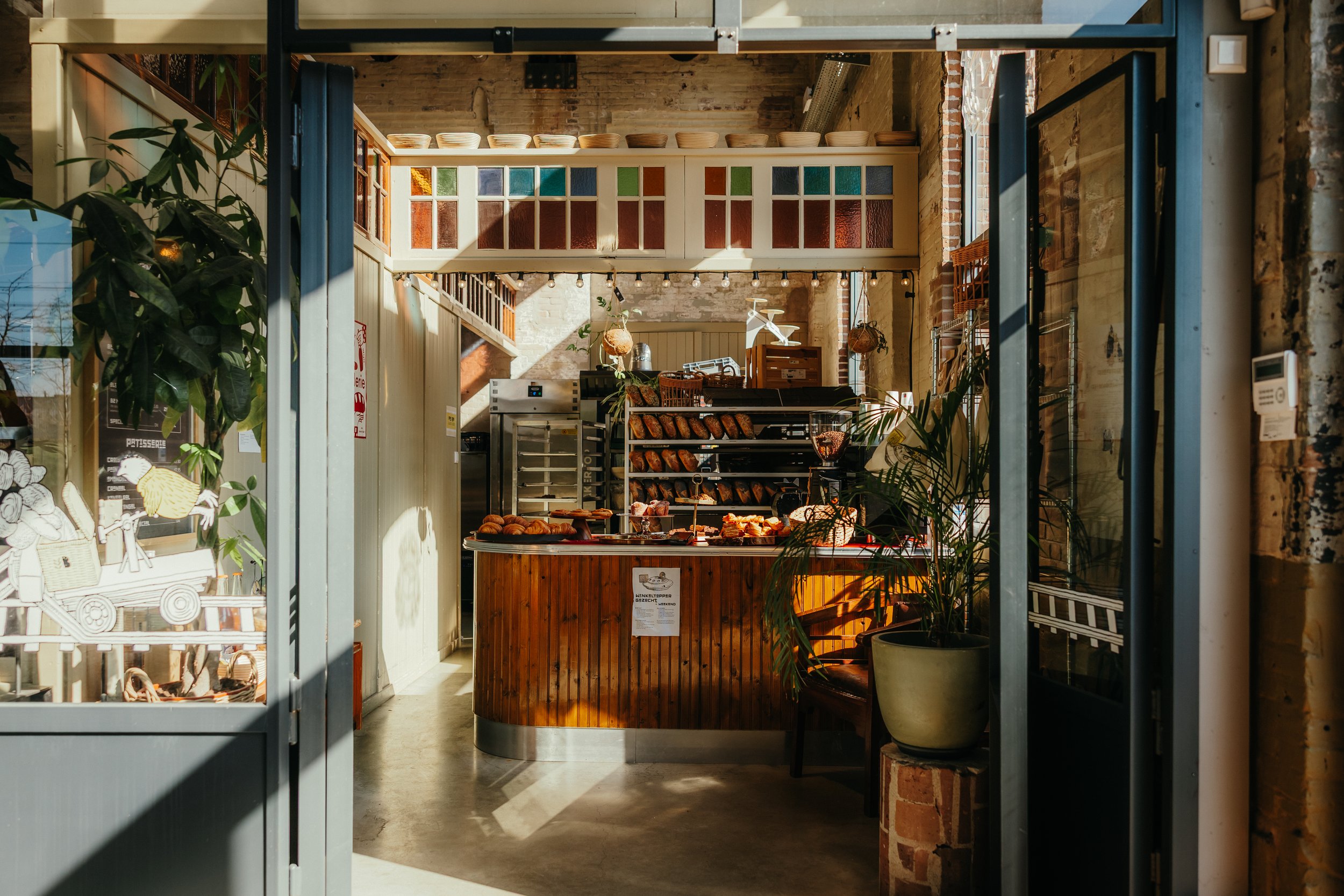 Interieur van een bakkerij of delicatessenwinkel met een toonbank vol gebak en brood, gekleurde glasramen boven de toonbank, planten en houten elementen, veel natuurlijk licht.