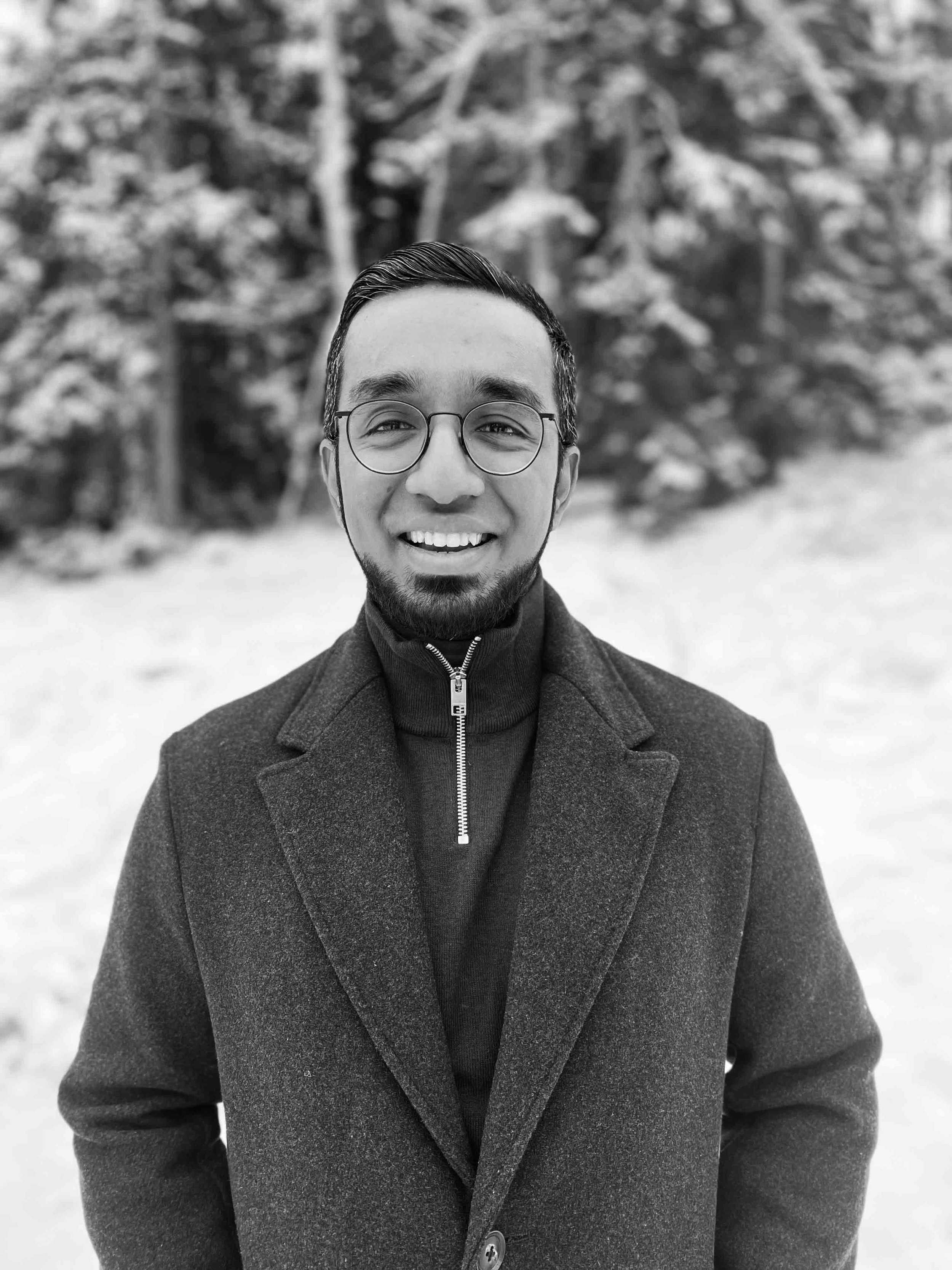 A smiling man with glasses and a beard wearing a dark coat and a zip-up sweater, standing outdoors in front of blurred trees.