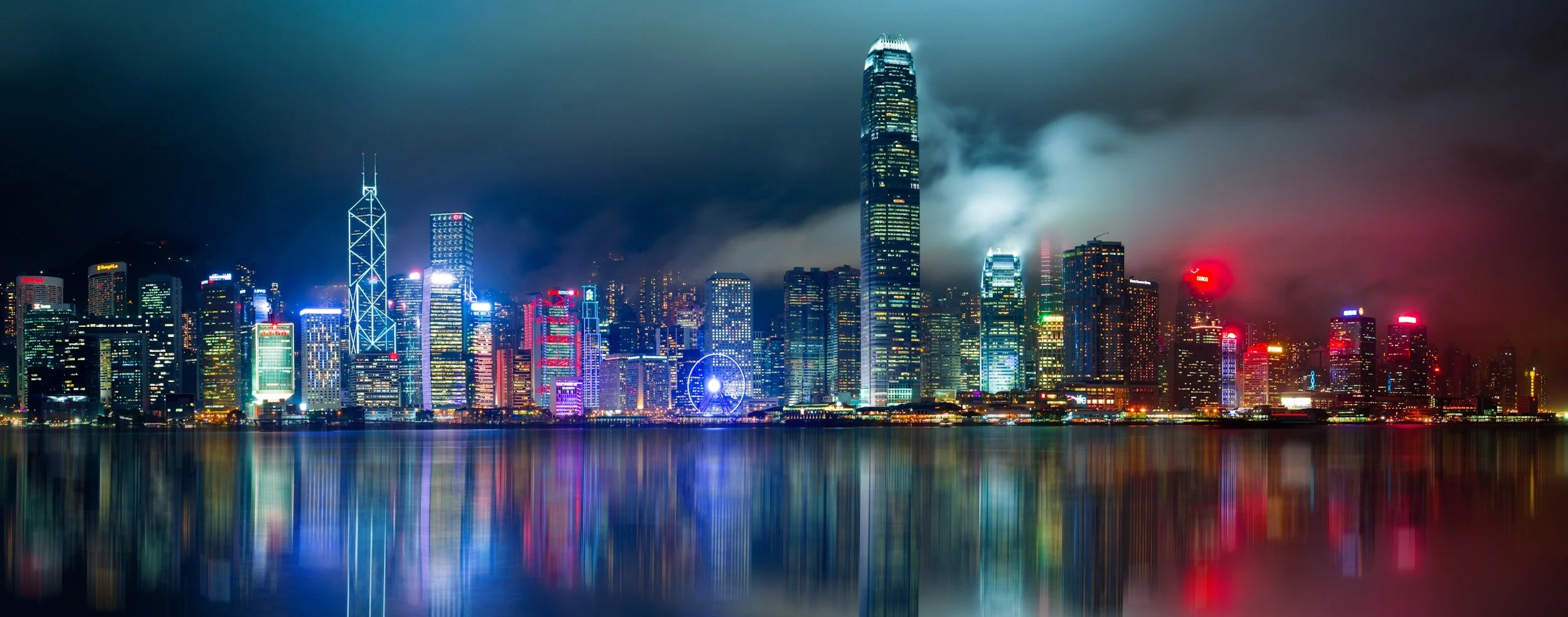 Nighttime city skyline with illuminated skyscrapers reflecting on water, with dark clouds overhead.