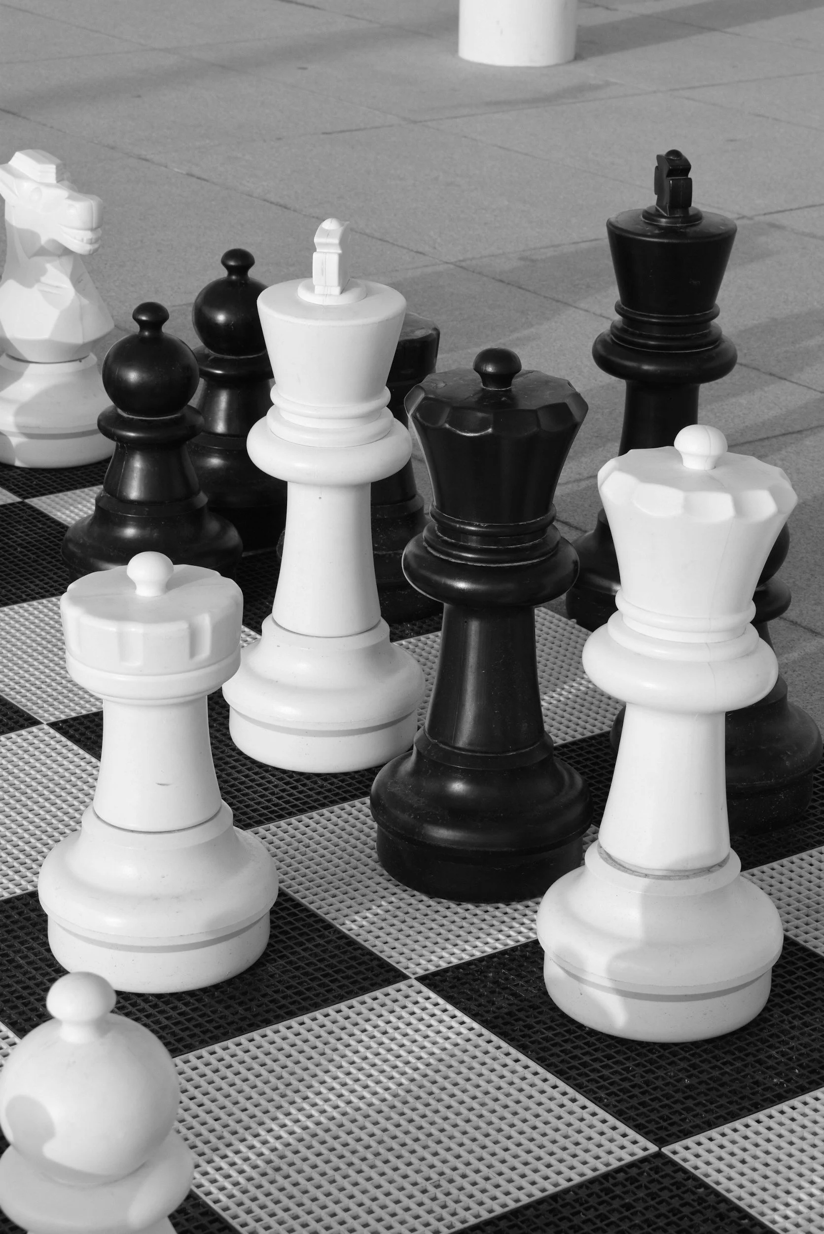 Black and white photo of large chess pieces set up on an outdoor chessboard, with some pieces on a raised platform, corner of the chessboard visible.