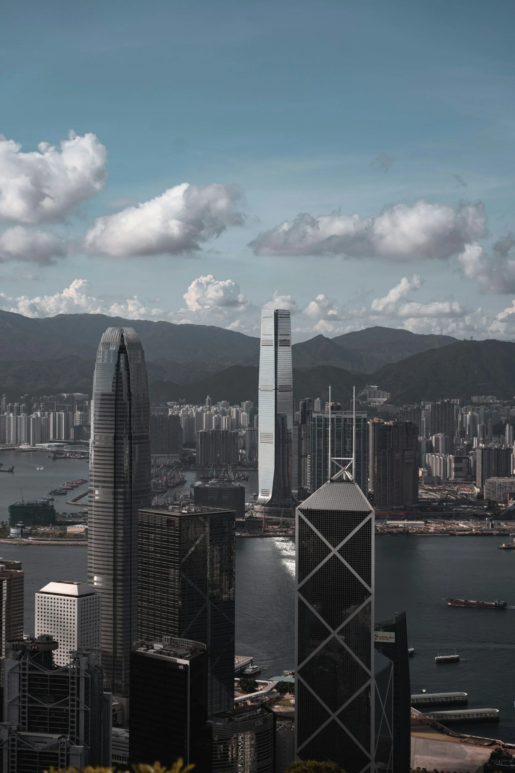 A cityscape featuring tall skyscrapers along a harbor with boats, mountains in the background, and a partly cloudy sky.