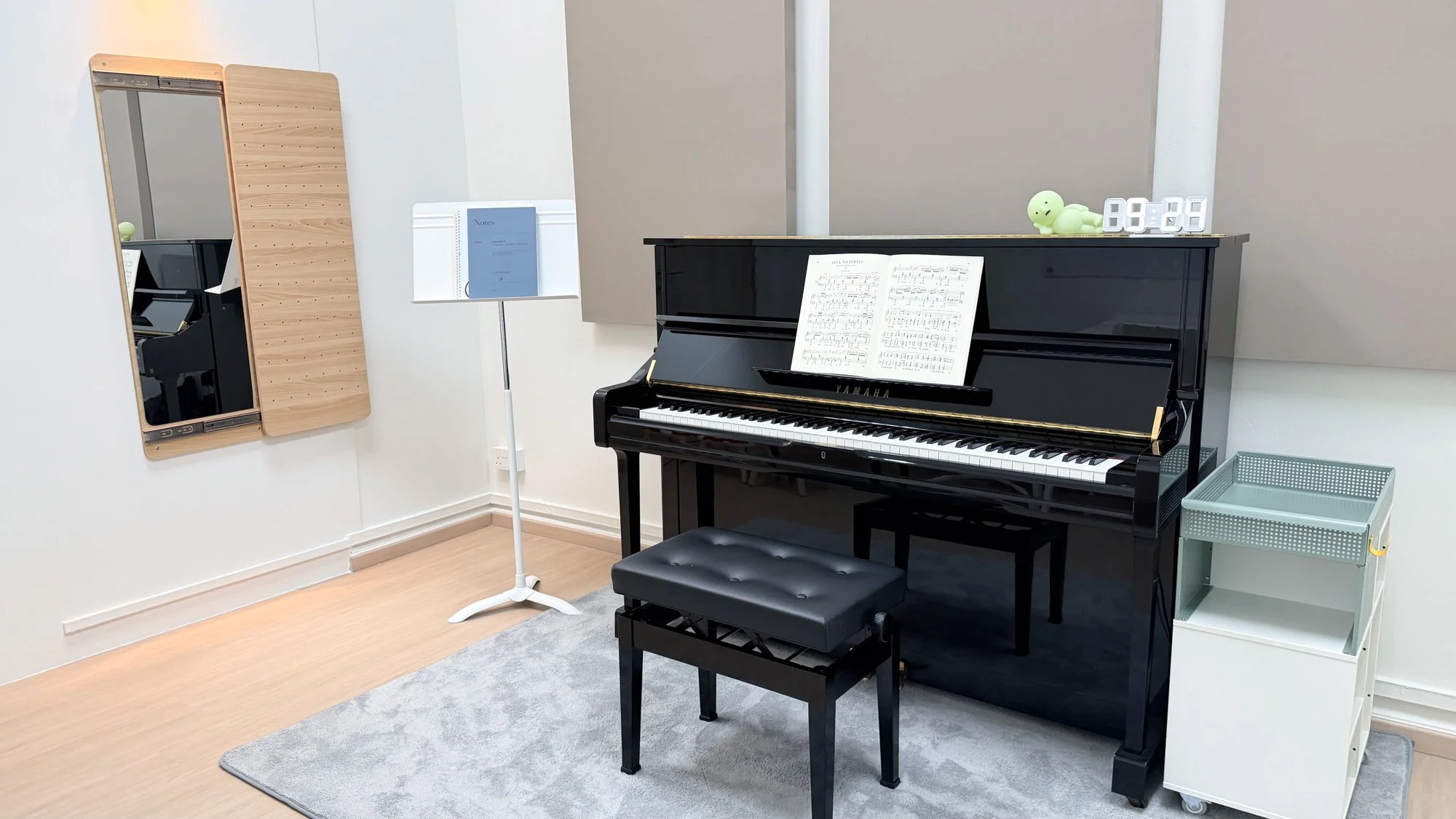 A black upright Yamaha piano positioned next to a green cushioned chair with floral pillows, big potted plant, and curtains by a glass door in a bright living room.