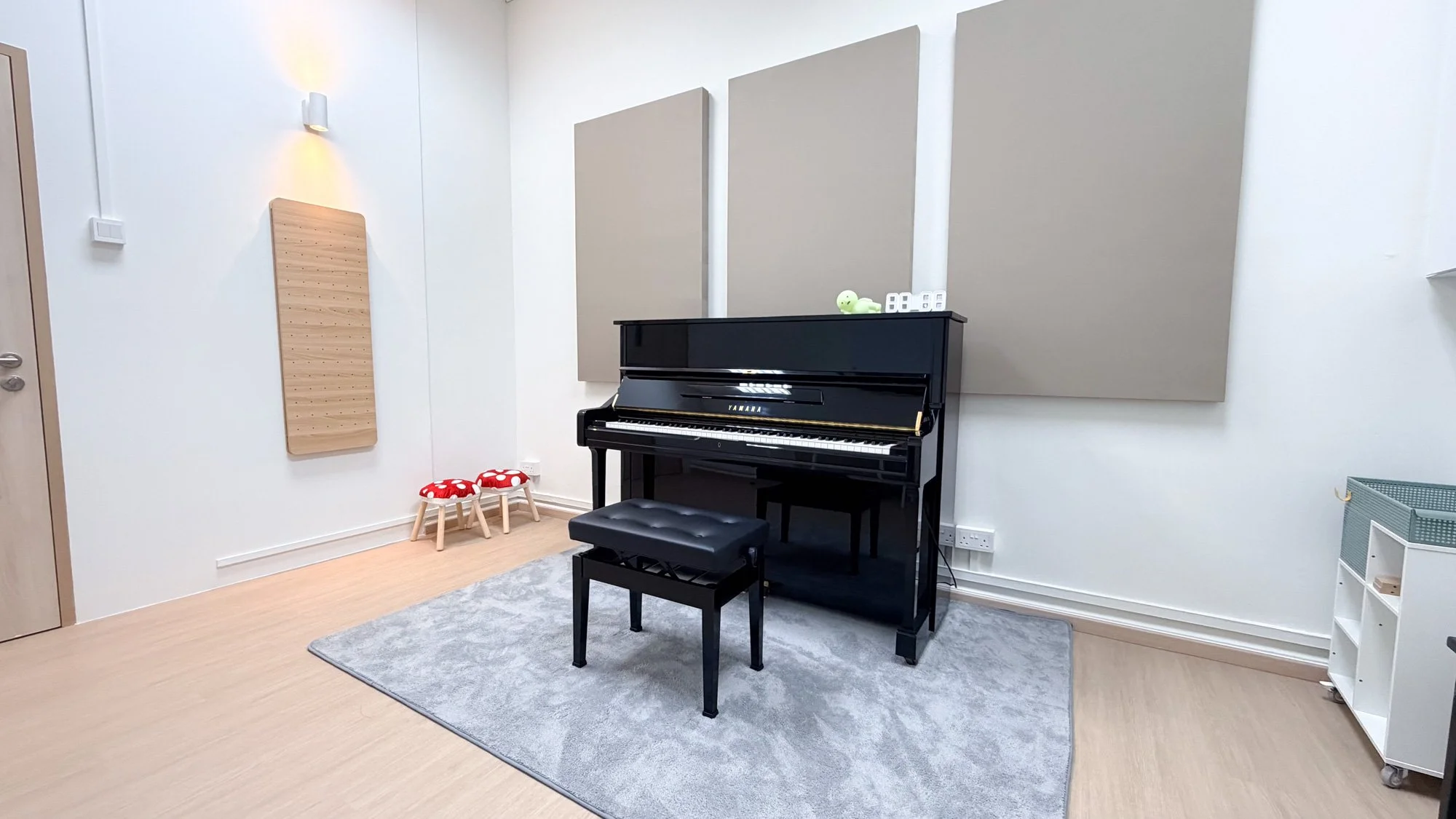 A black upright Yamaha piano positioned next to a green cushioned chair with floral pillows, big potted plant, and curtains by a glass door in a bright living room.