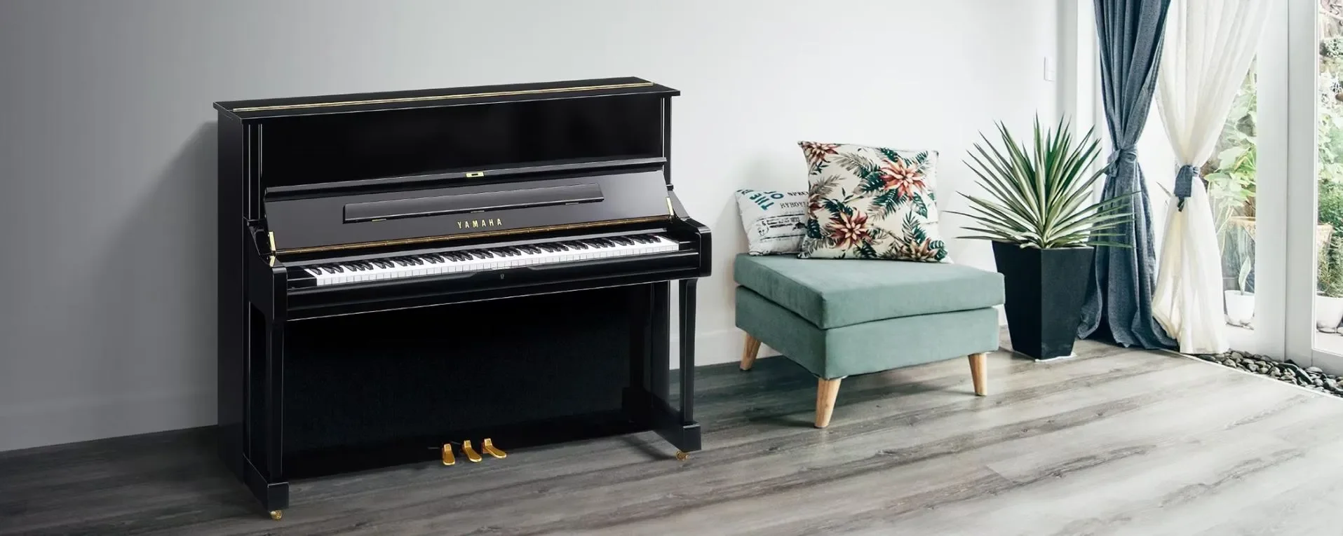 A black upright piano with gold accents next to a teal armchair with floral and log cabin patterned pillows, positioned near a large window with blue and white curtains. There is a tall potted plant with long narrow leaves beside the window, and a view of lush greenery outside.