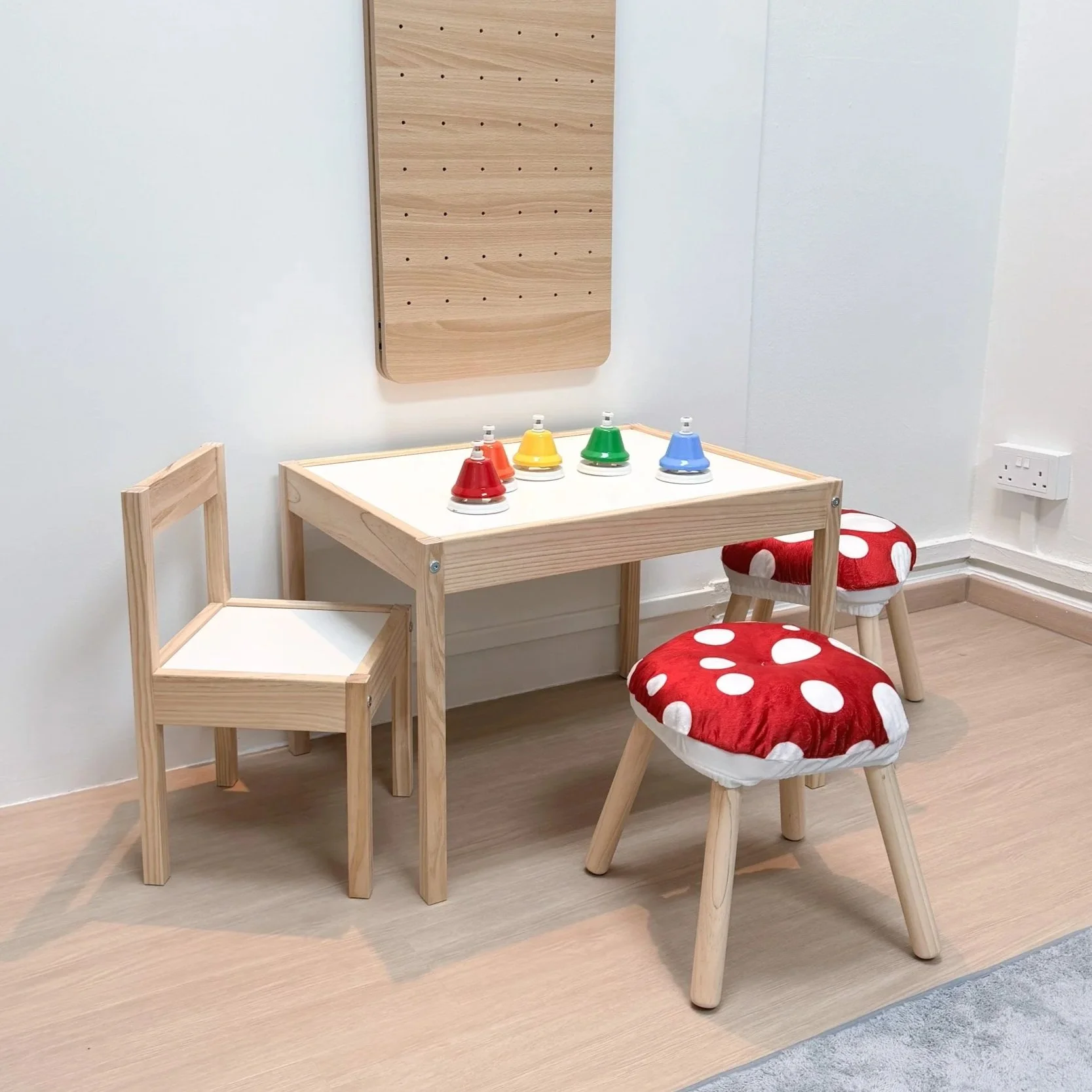 A children's play area with a small wooden table and chairs, colorful bell stacking toys on the table, a stool with a mushroom-print cushion, and a wooden wall panel in a room with white walls and light wood flooring.