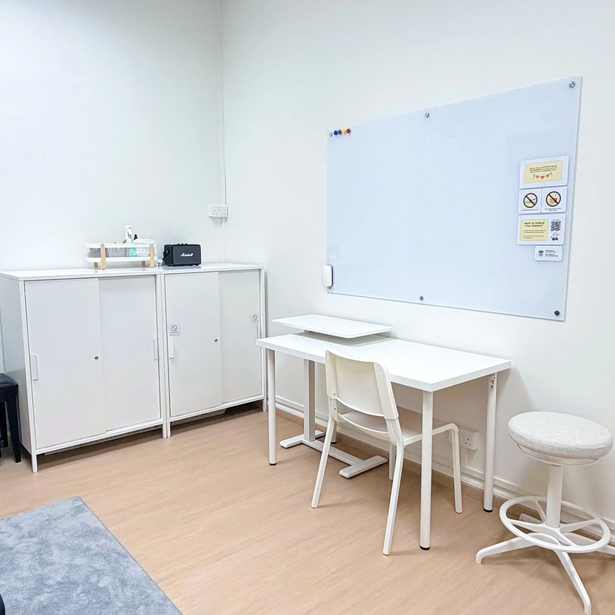 A white room with a whiteboard on the wall, a white table with two white chairs, and a white cabinet with a small speaker, a basket, and some items on top. The floor is a light wood color with blue and gray rugs.