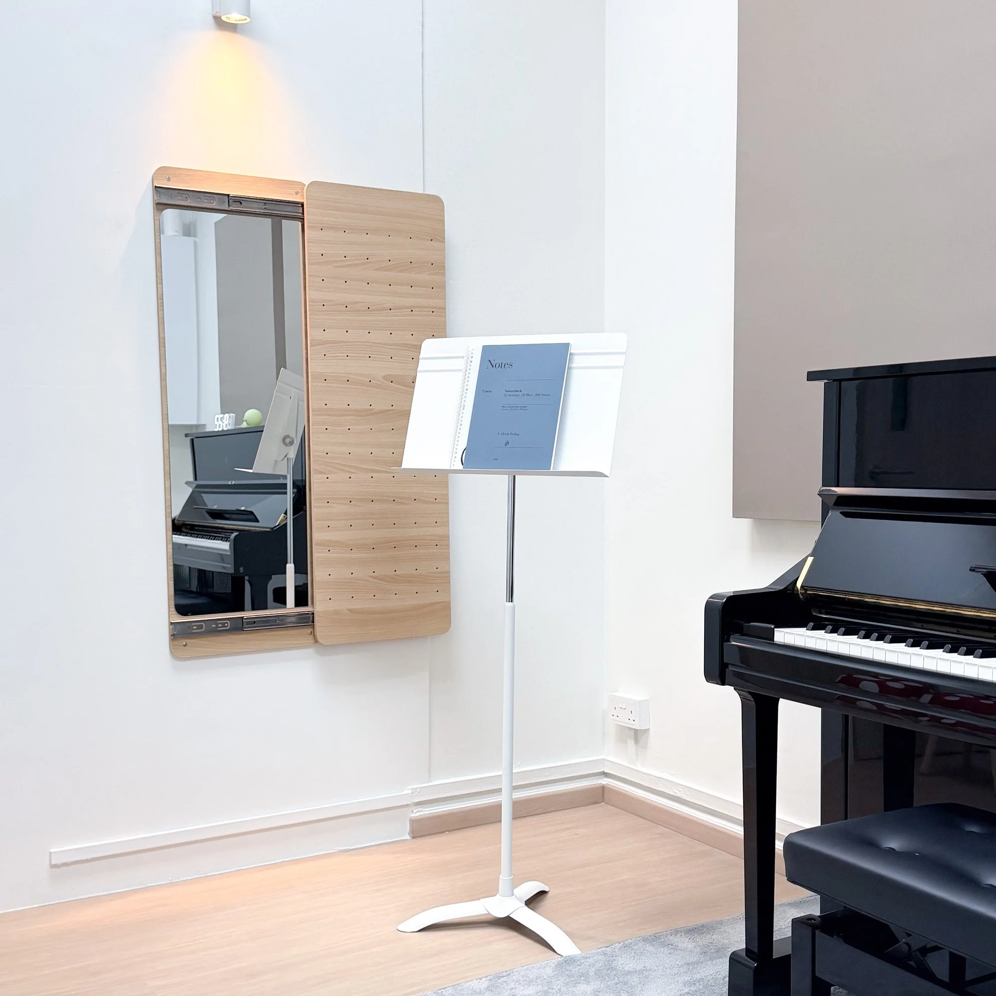 A minimalistic room with a black upright piano centered against a plain white wall, a small matching black stool in front, a large window with light streaming in on the right, a brown rug on light wood flooring, and a built-in wooden bookshelf filled with books on the left wall.