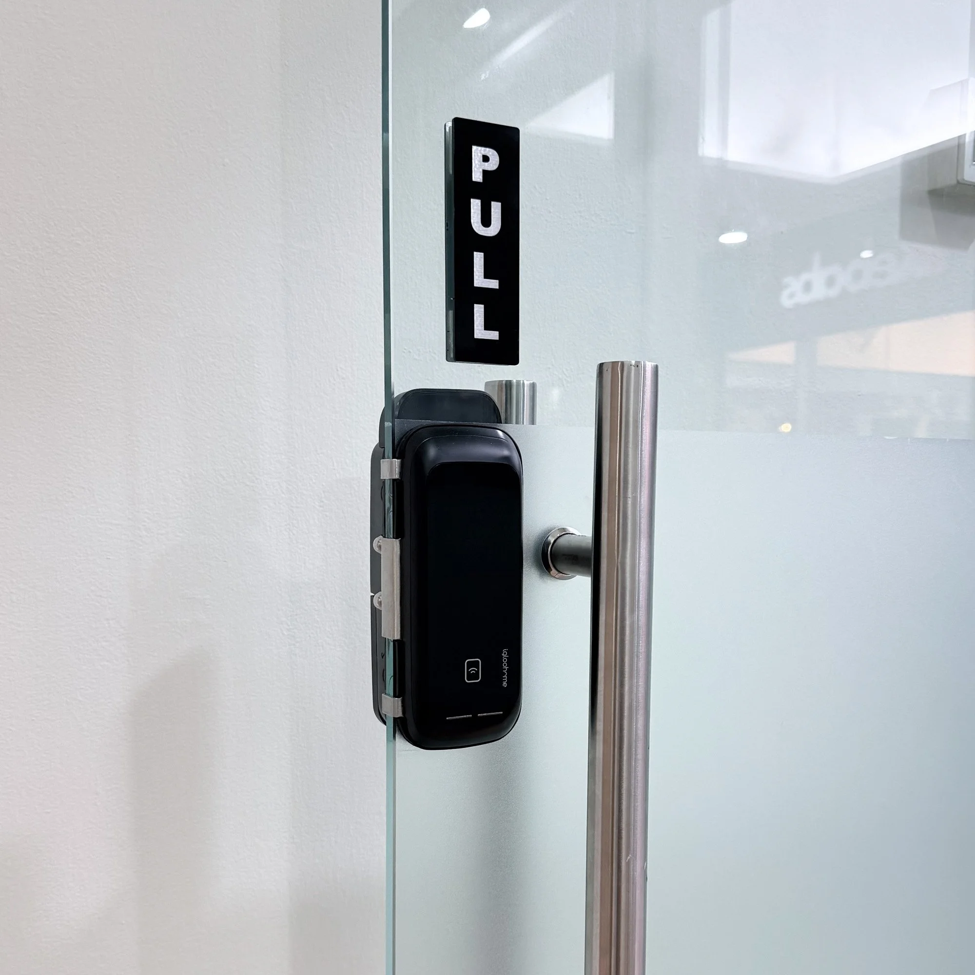 A close-up view of a black smart lock with a numeric keypad, mounted on a door in an office setting. In the background, there is a blurred office chair and a table. The image is split with a visible seam down the middle.