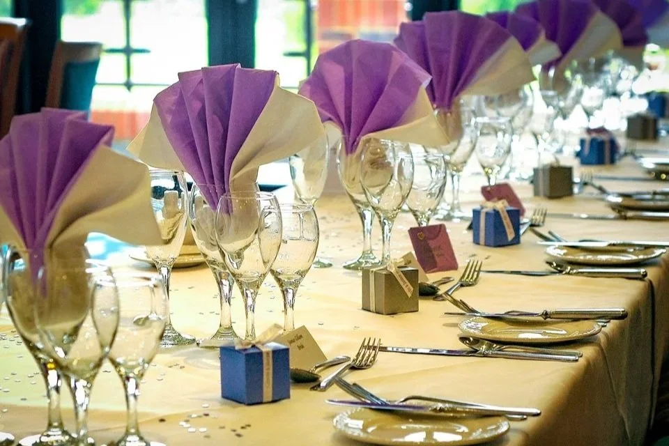 A long banquet table set with wine glasses, folded purple and beige napkins, small gift boxes, plates, silverware, and name cards.