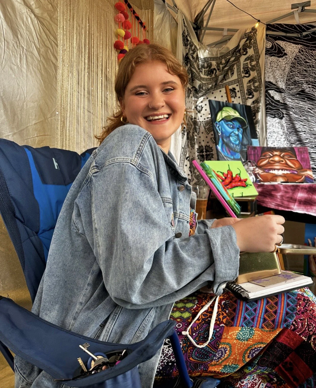 A young woman with red hair smiling at an art booth with colorful paintings and handmade items around her.