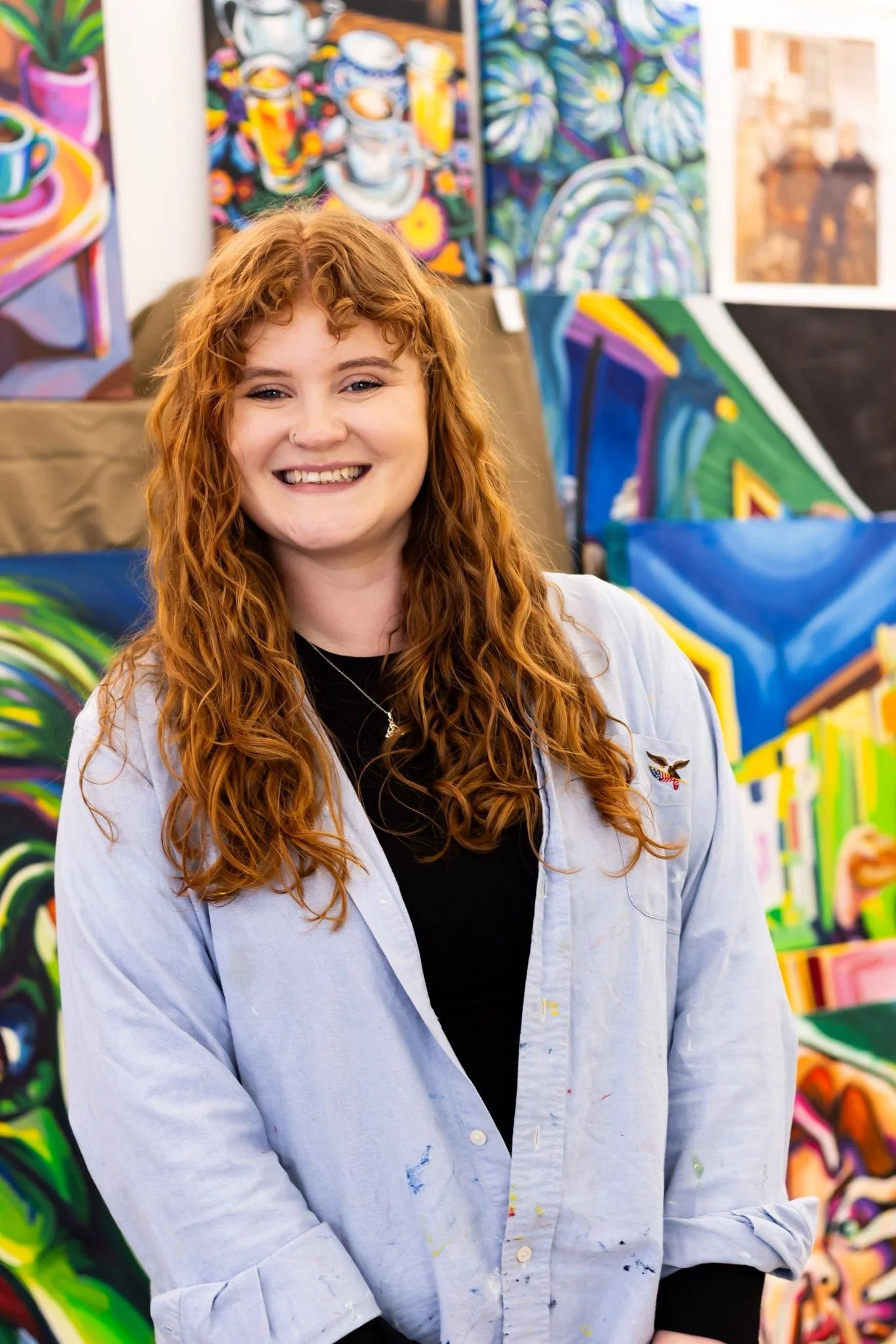 Young woman with long, curly red hair smiling in art studio with colorful paintings in the background.