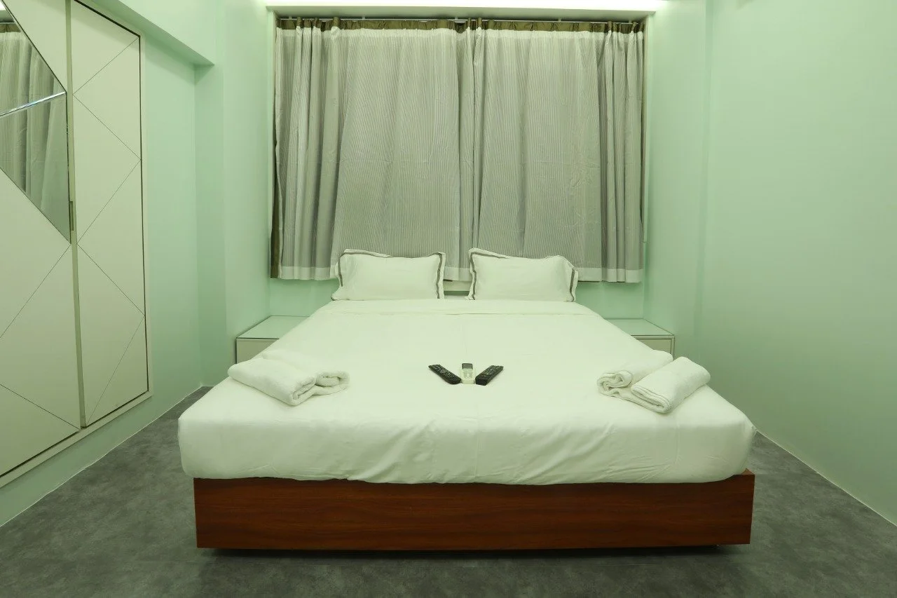 A minimalist hotel room with a bed, two pillows, and two rolled towels on the bed. There are two remote controls on the bed, and a window with closed curtains behind it. The room has light-colored walls and a carpeted floor.