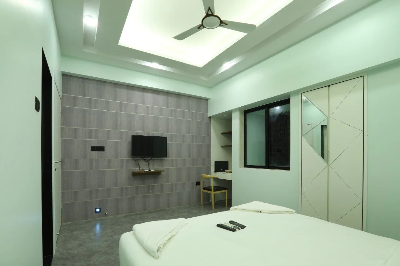 Quiet hotel room with a bed, two remote controls, a TV on the wall, a window, a small desk with a chair, and modern decor.