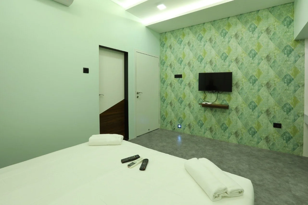 Modern hotel room with a bed, remote controls, towels, TV mounted on a green patterned accent wall, and minimalist decor.